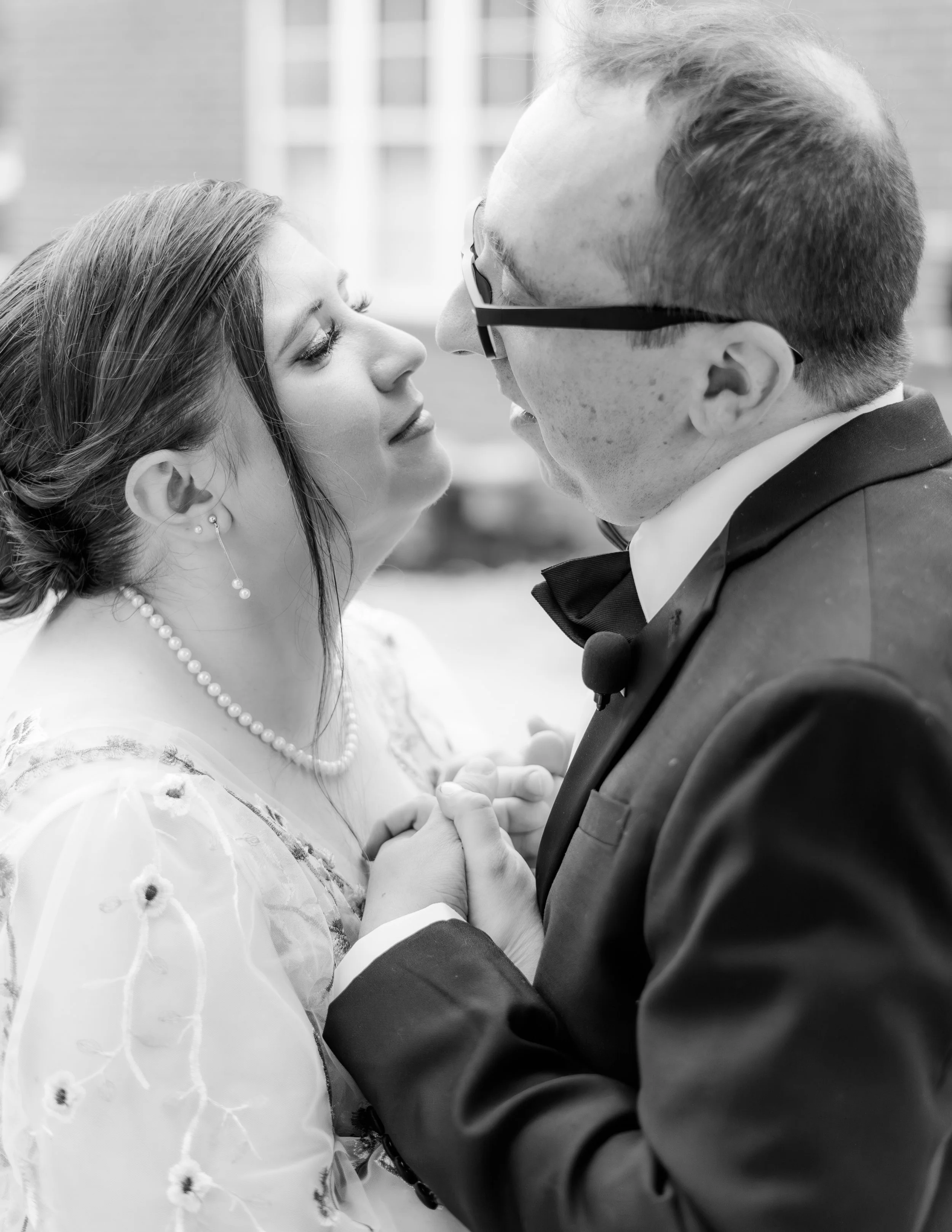 A black and white photo of a bride and groom close together, holding hands, about to kiss during their wedding.