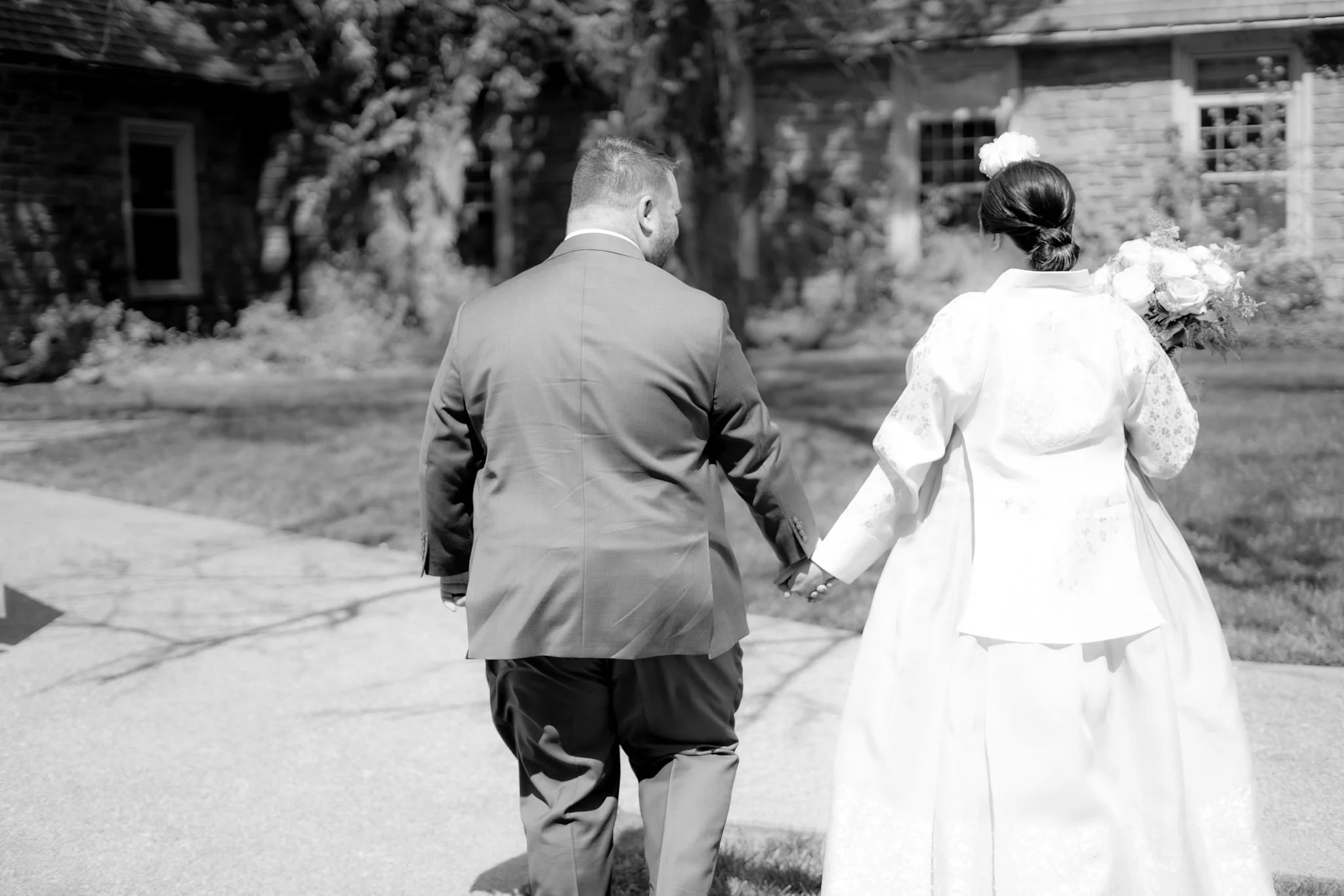 A black and white photo of a man and woman holding hands, walking away from the camera outdoors, possibly during a wedding or romantic outing. The woman is wearing a white dress and holding a bouquet of flowers, while the man is dressed in a suit. Th