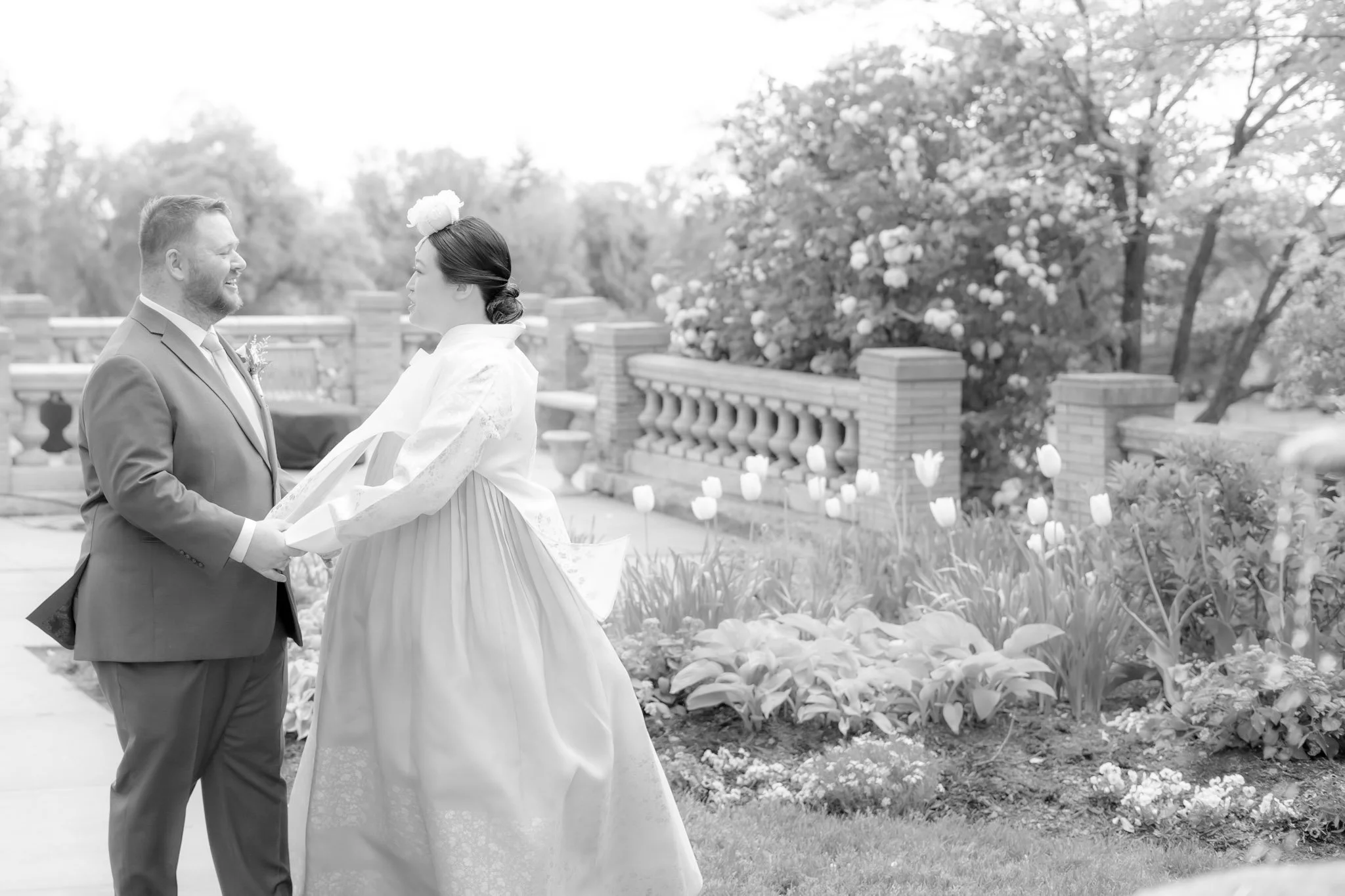 A couple dressed in wedding attire holding hands and looking at each other outdoors, with flowering trees and bushes in the background.