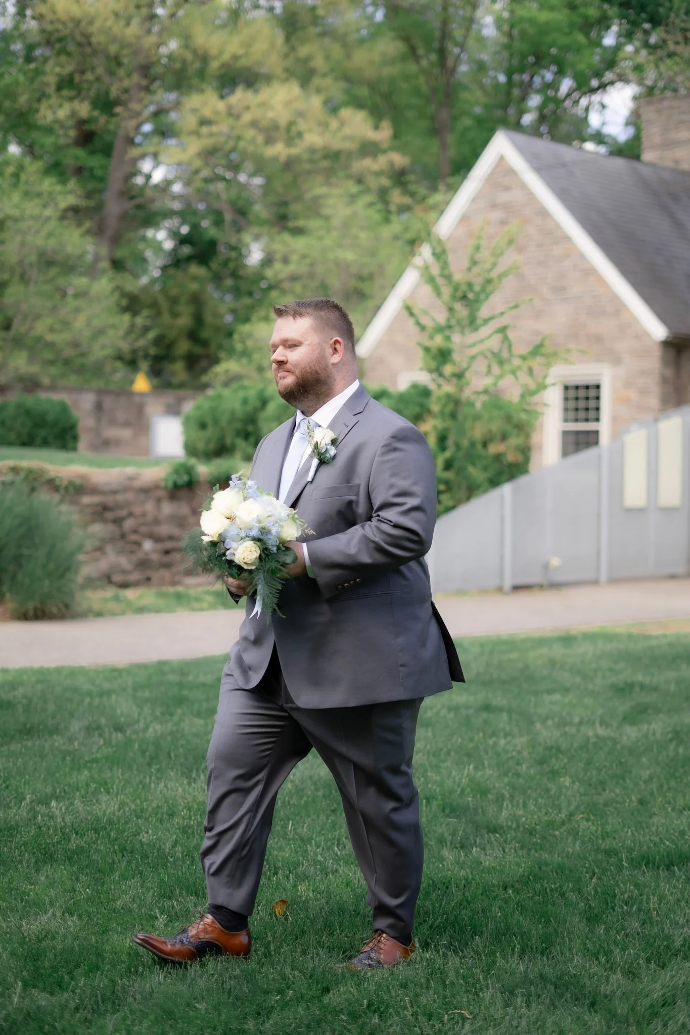 A man in formal attire holding a bouquet of flowers outdoors, with a house and trees in the background.