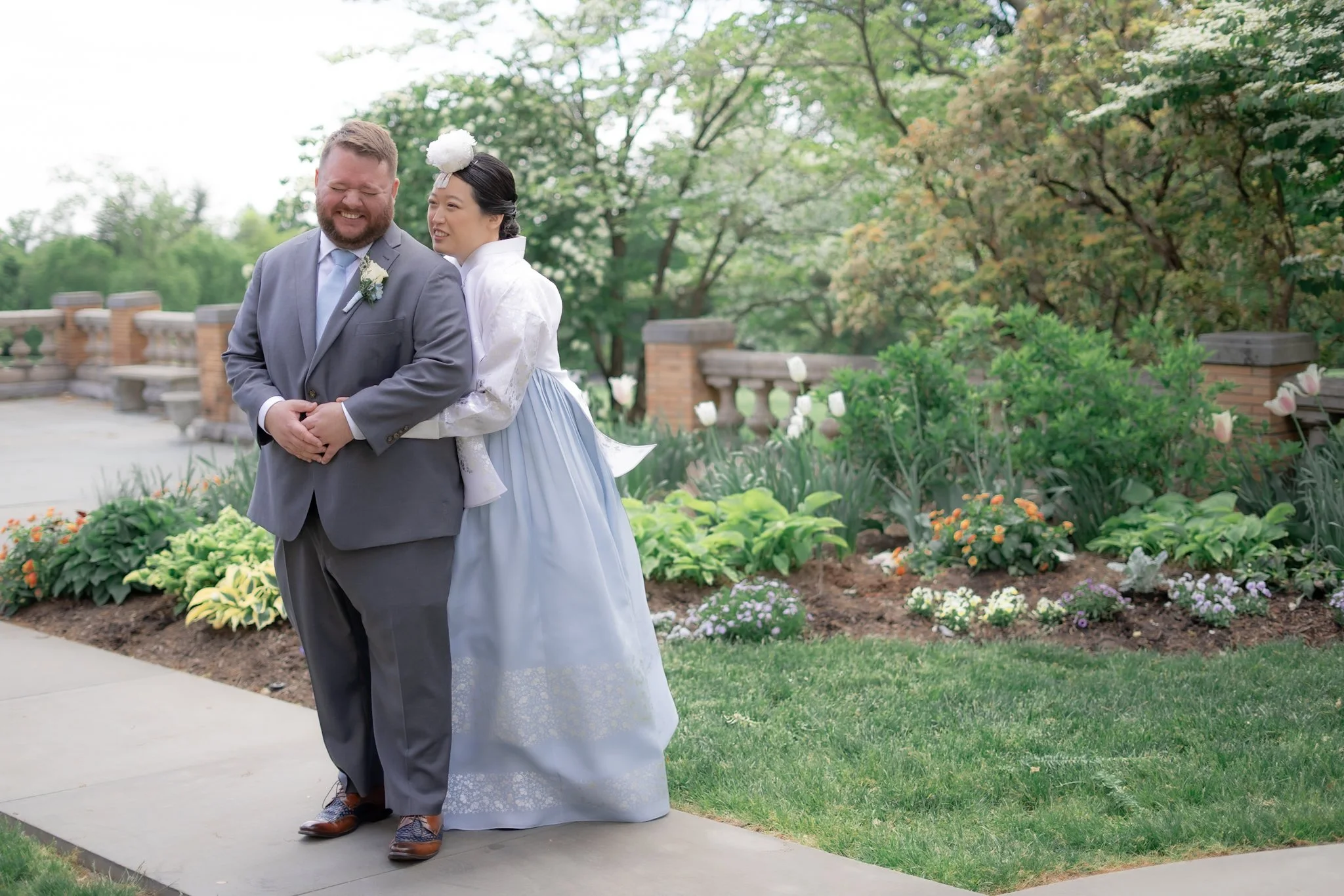 A newlywed couple standing outdoors on a sidewalk near a garden with colorful flowers and green trees. The groom is wearing a gray suit, and the bride is in a traditional white wedding dress with a white floral headpiece. They are smiling and embraci