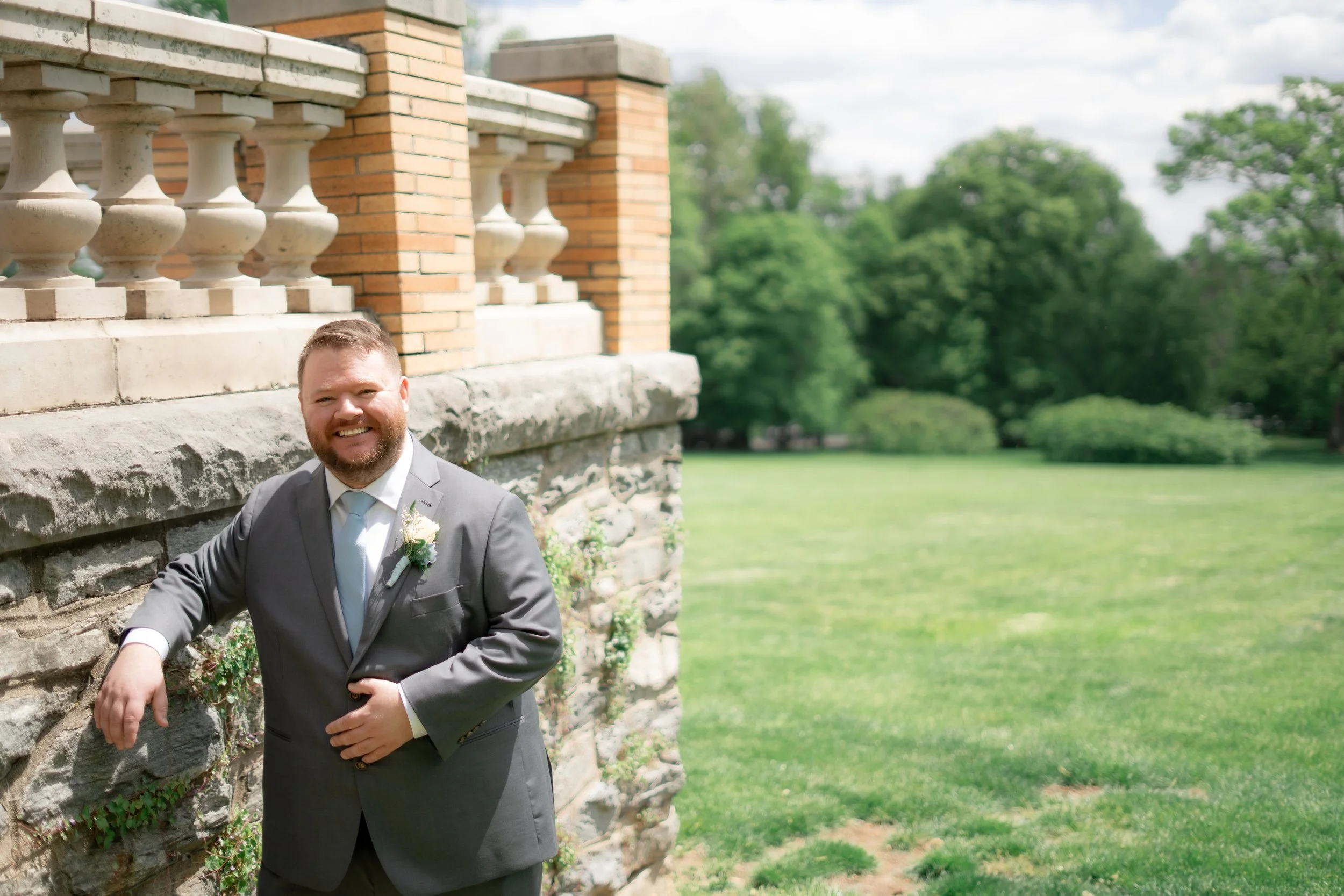 A smiling man in a gray suit with a boutonniere, standing outdoors near a stone and brick structure with a lush green background and trees.