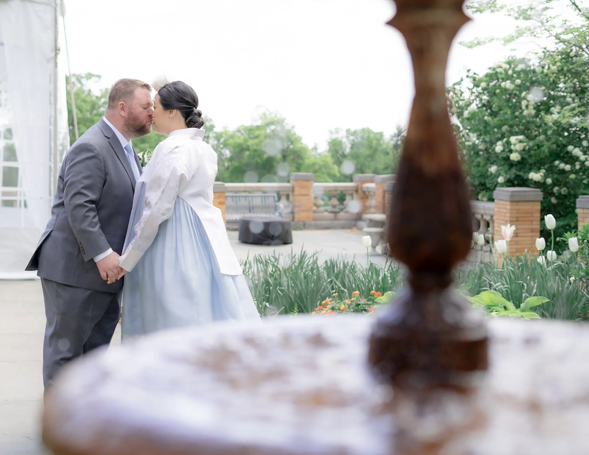 A couple in wedding attire sharing a kiss on an outdoor terrace, with blurred garden scenery and a wooden element in the foreground.