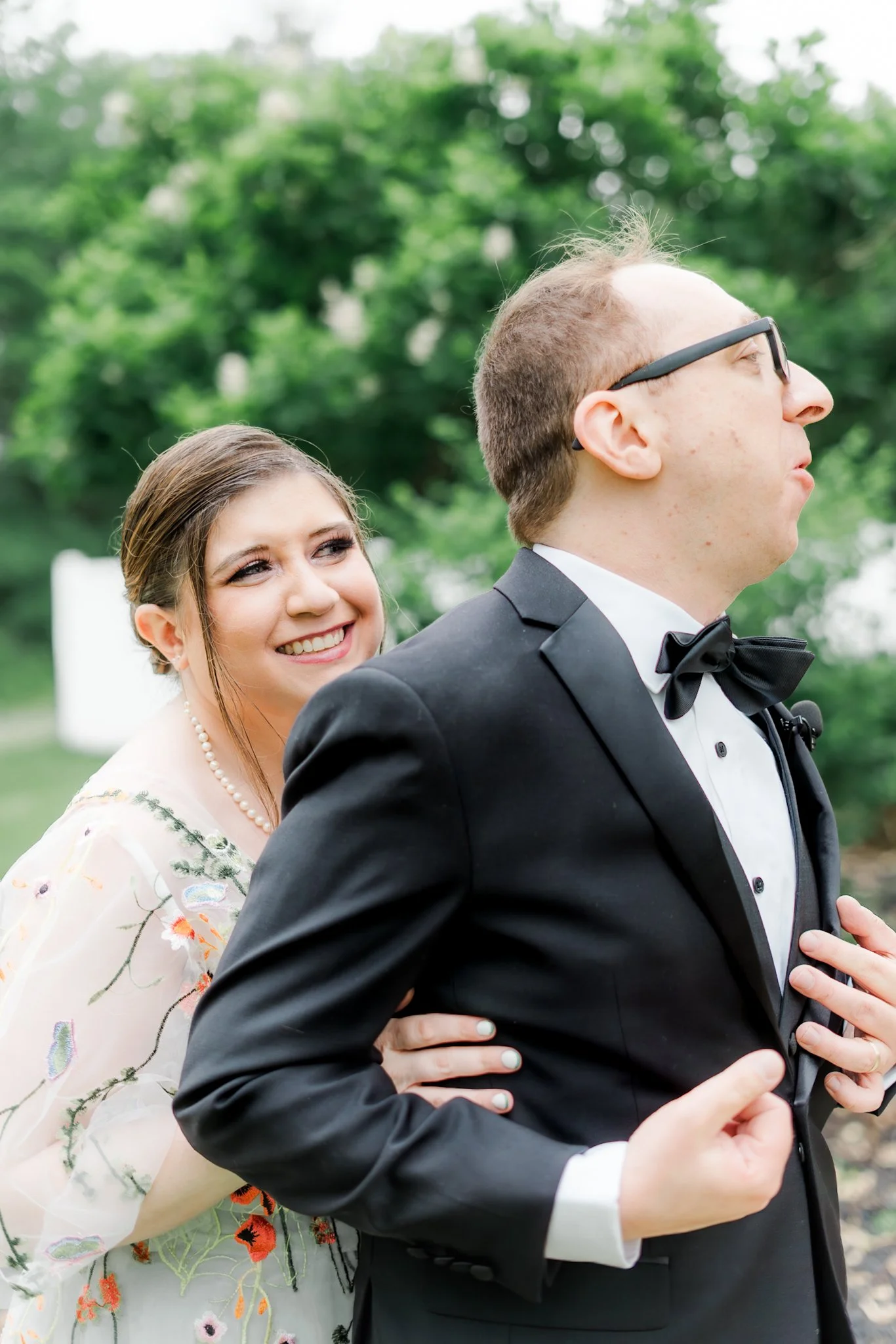 A bride smiling behind a groom wearing glasses and a tuxedo, with his hand on his chest, outdoors with green blurred trees in the background.