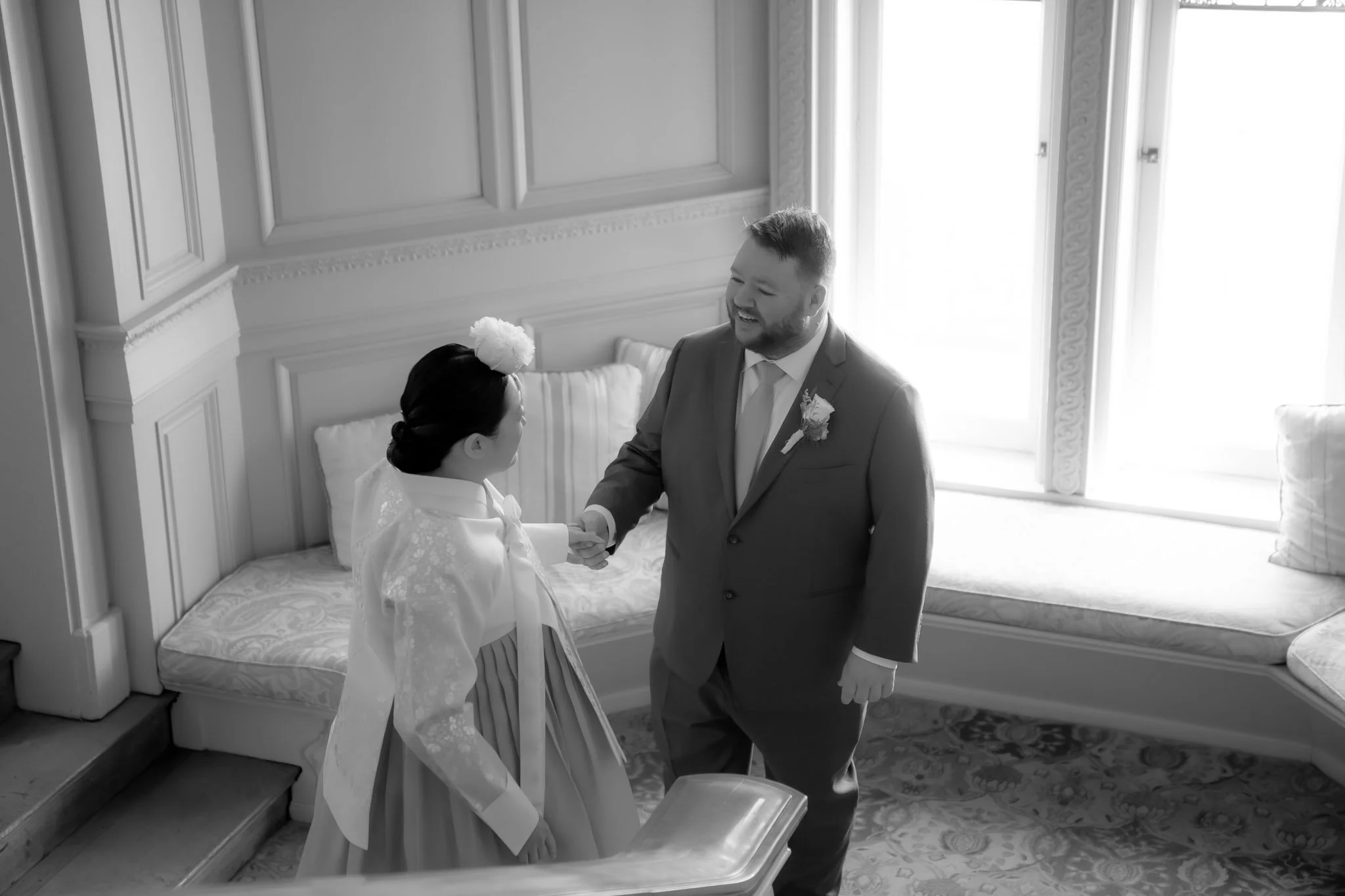 A bride and groom holding hands and smiling at each other inside a bright room with large windows and ornate woodwork. The bride wears a white dress with lace details and a flower headpiece, while the groom wears a suit with a boutonniere.