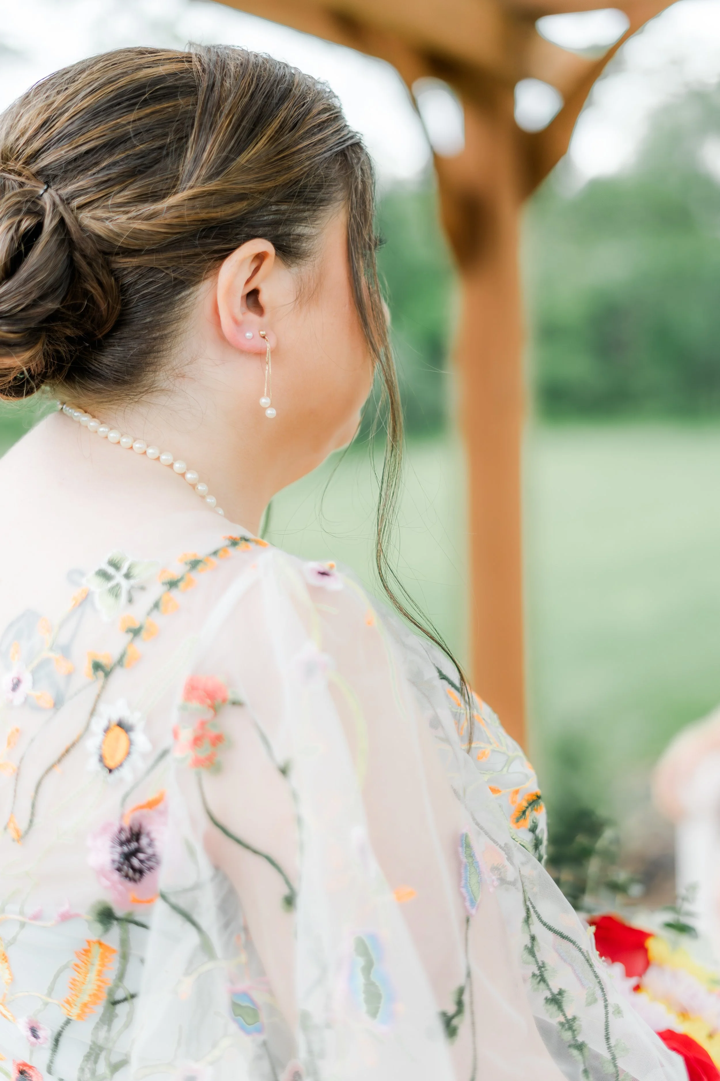 A woman with brown hair styled in an updo, wearing pearl earrings and a pearl necklace, is seen from the side in a blurry outdoor setting with wooden structures and greenery.