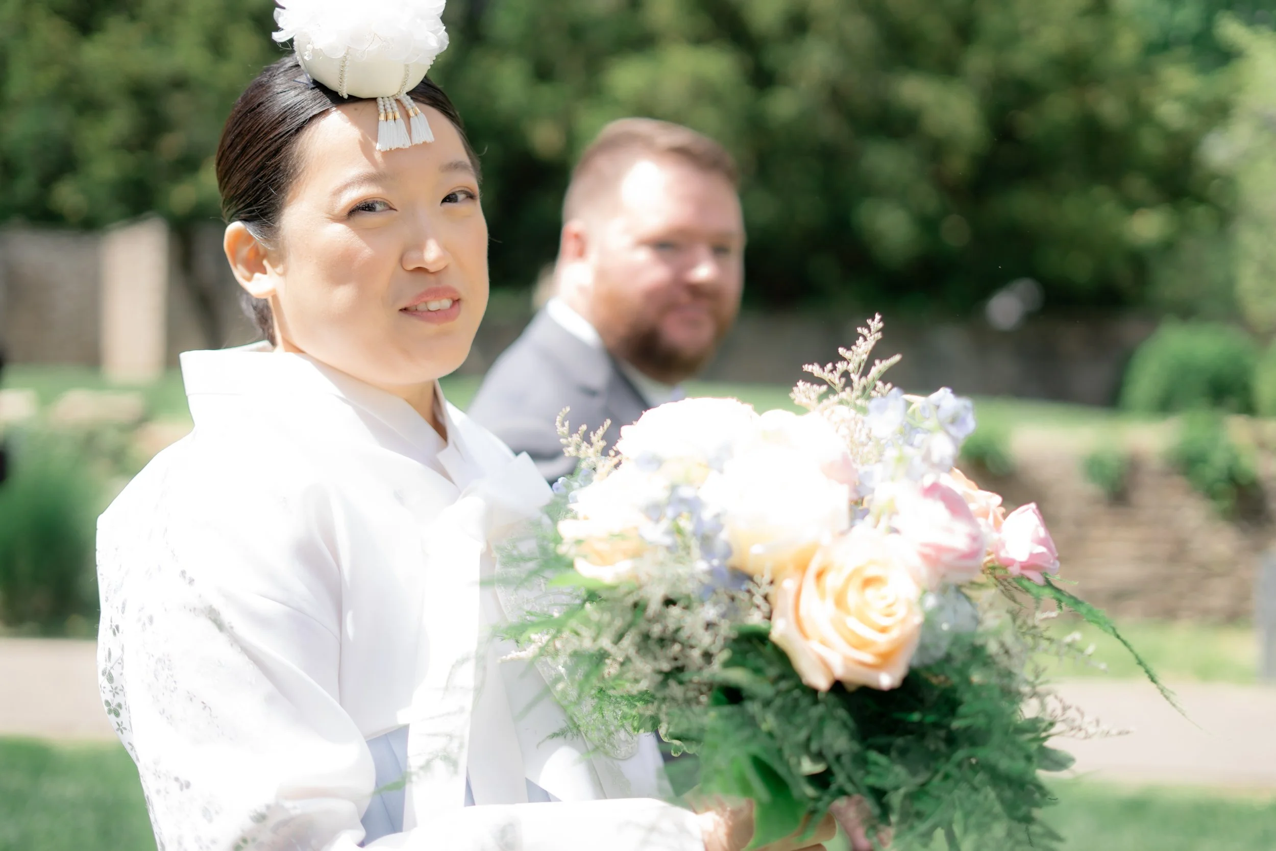 A woman dressed in traditional Asian attire holding a bouquet of pastel-colored flowers, with a man in a gray suit standing behind her outdoors on a sunny day.