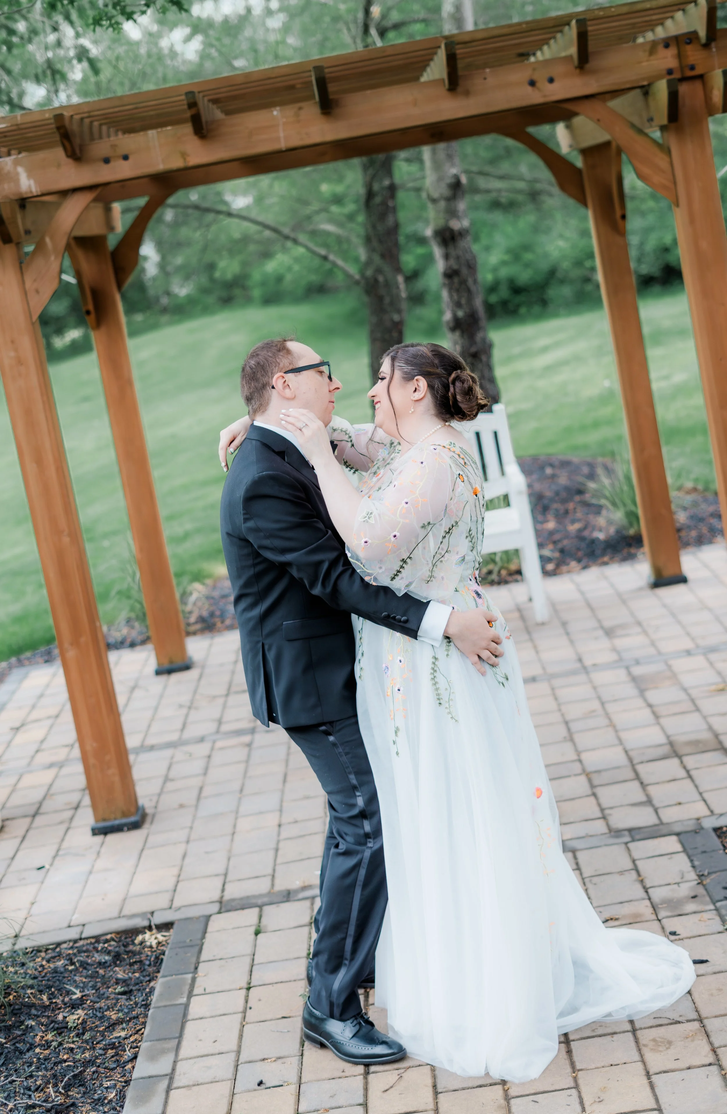 A bride and groom dance outdoors under a wooden pergola with a green grassy background.