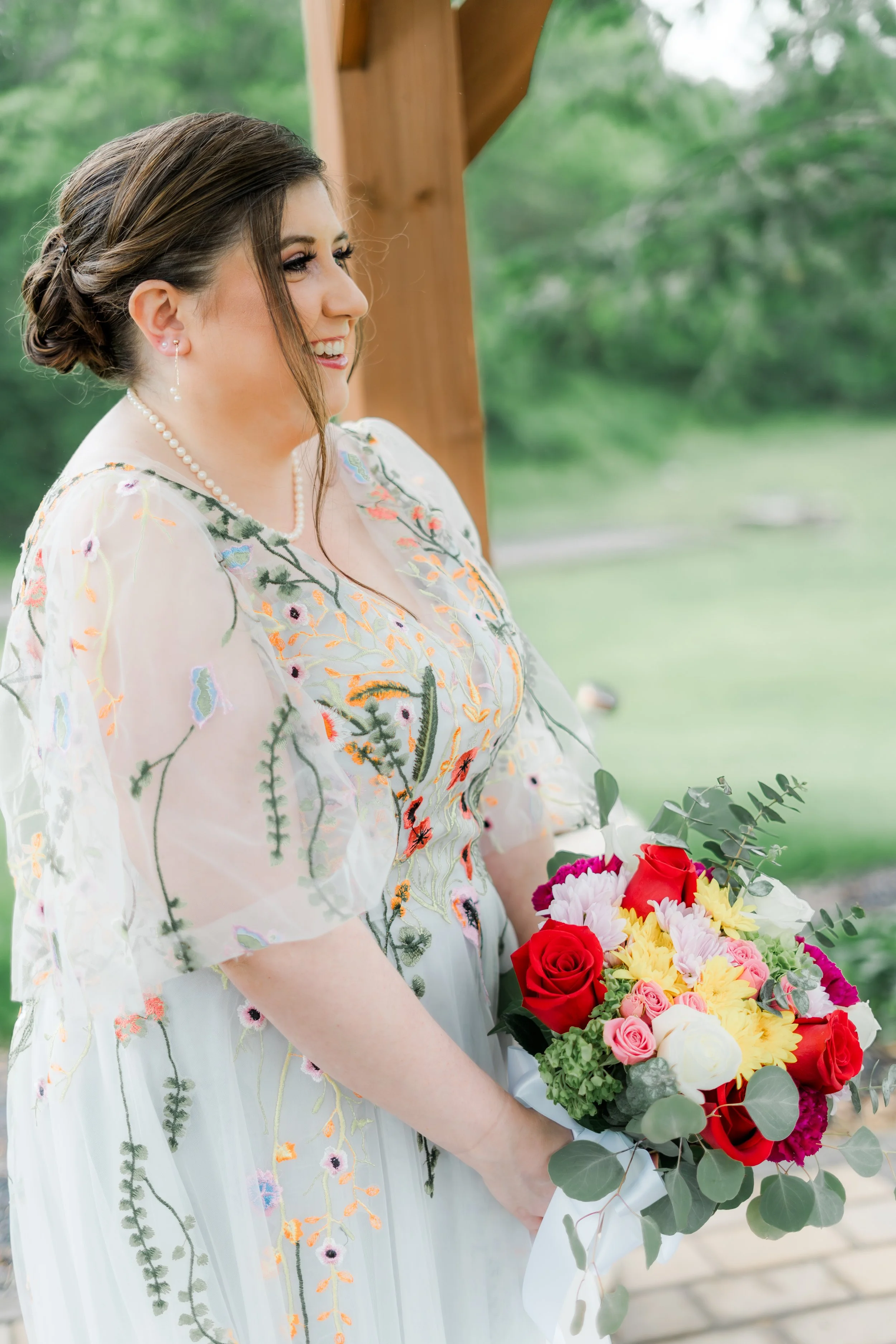 A woman dressed in a floral embroidery dress holding a bouquet of red, pink, yellow, and white flowers outdoors with a blurred green background.