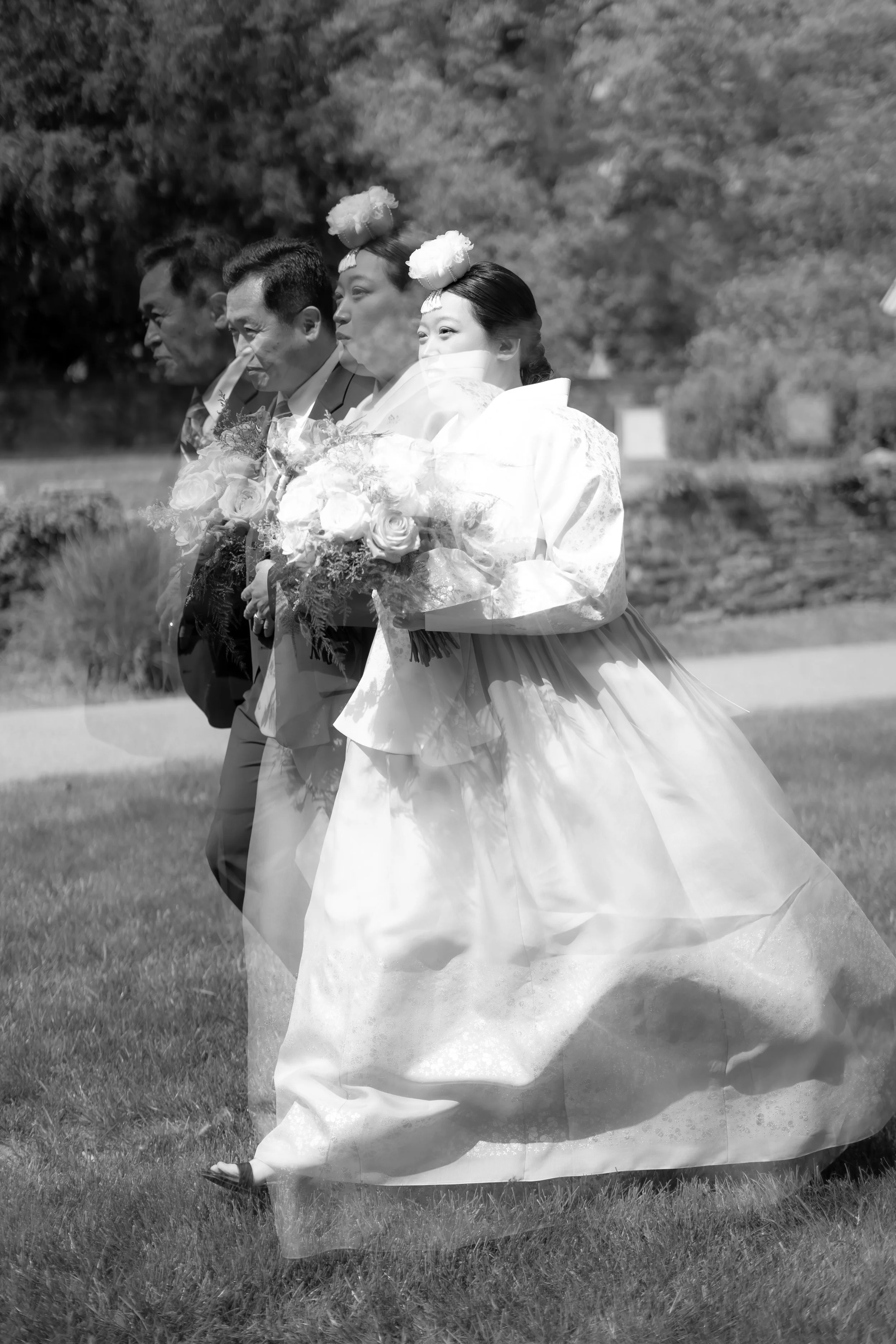 Group of five people in formal attire walking outdoors, holding bouquets, with trees and grass in the background, in a black and white photograph.