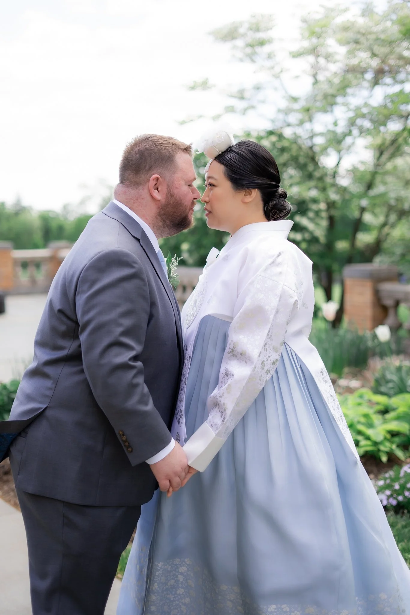 A couple holding hands outdoors with trees in the background, dressed in wedding attire, about to kiss.
