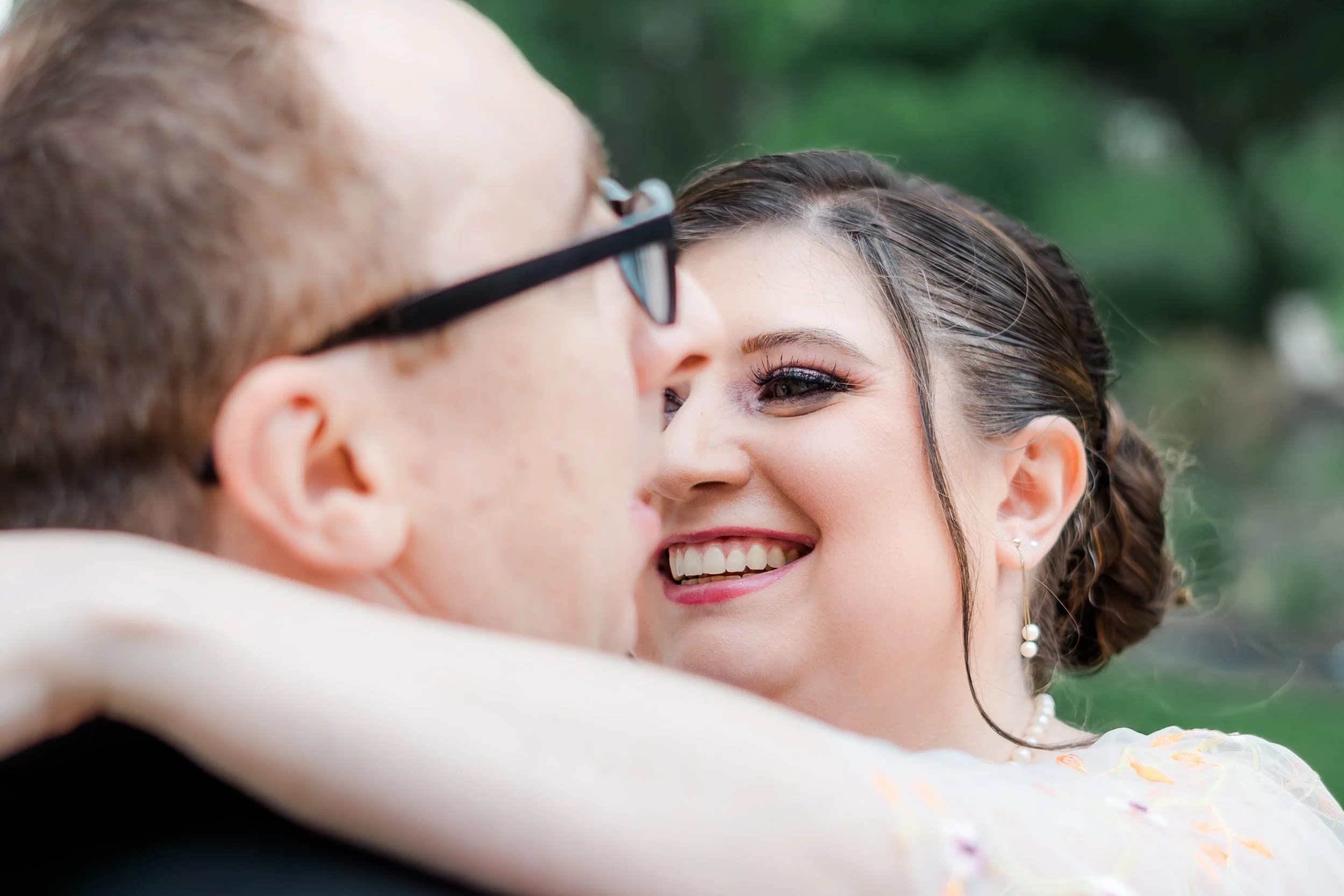 Close-up of a man and a woman embracing outdoors, with the woman smiling and her eyes visible, green blurred background.