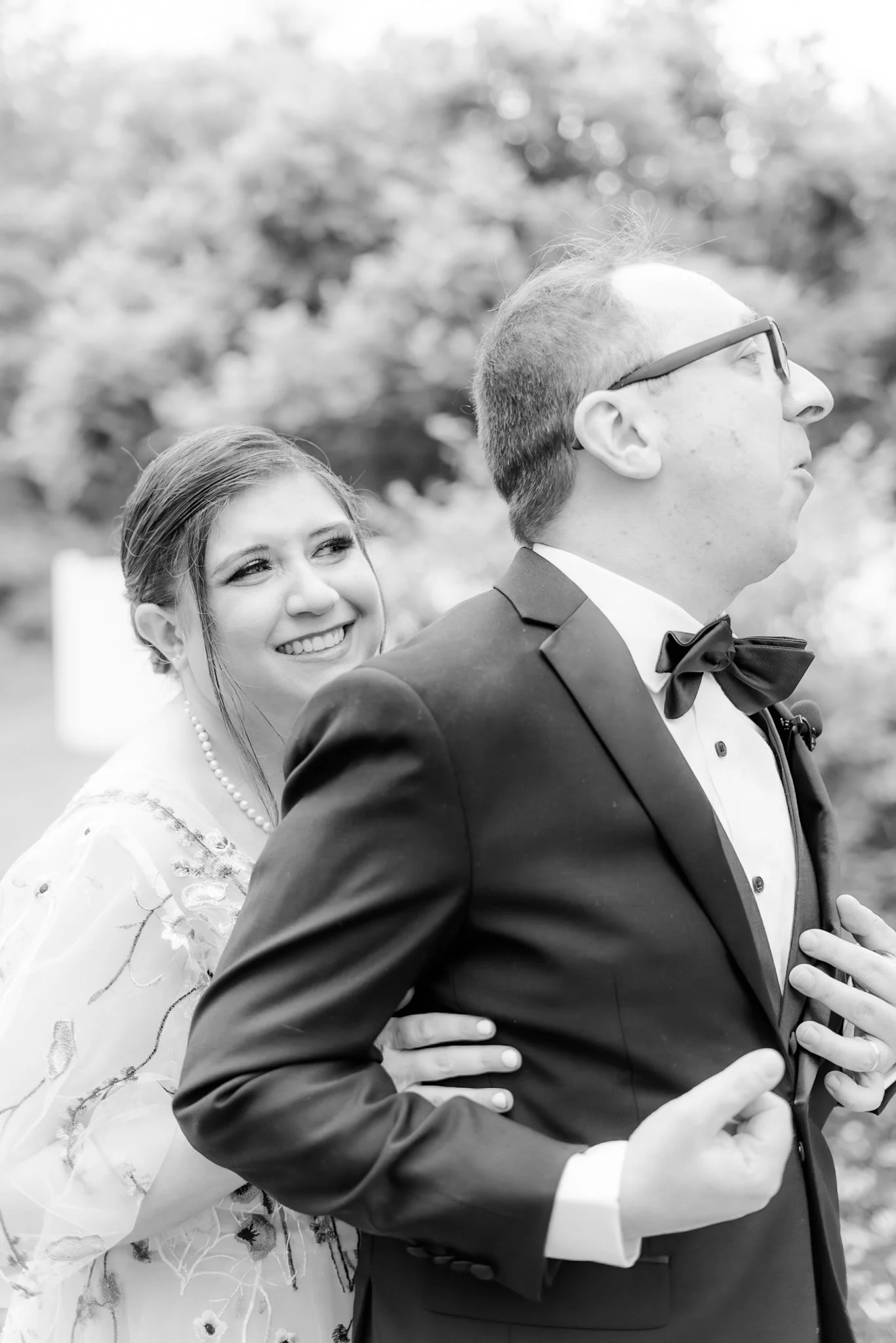 A black-and-white photo of a bride and groom, with the bride smiling and appearing happy, looking at the groom, who is dressed in a tuxedo with a bow tie, and wearing glasses, in an outdoor setting with blurred trees in the background.