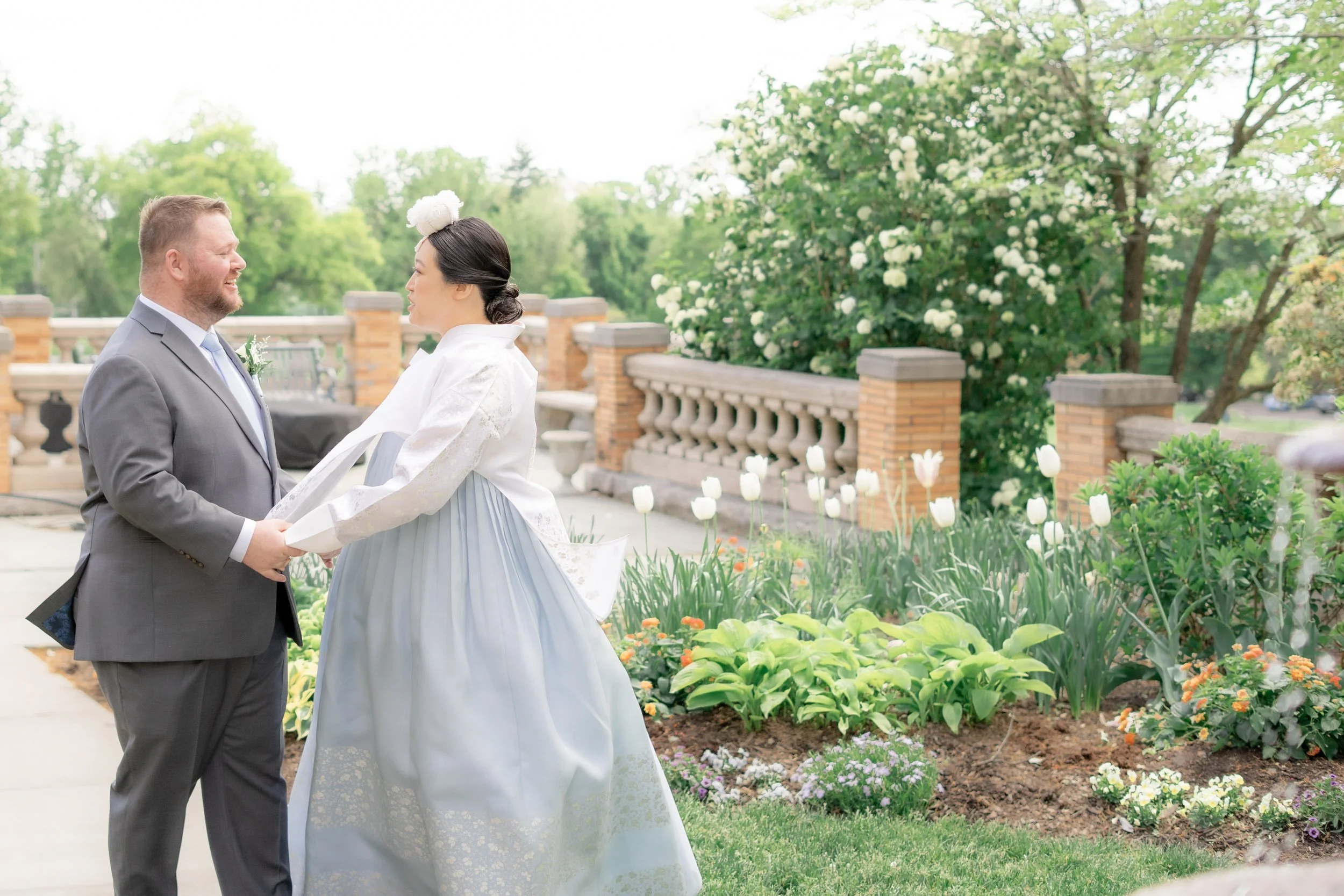 A bride and groom holding hands during their wedding ceremony outdoors in a garden with blooming white flowers and lush green trees.