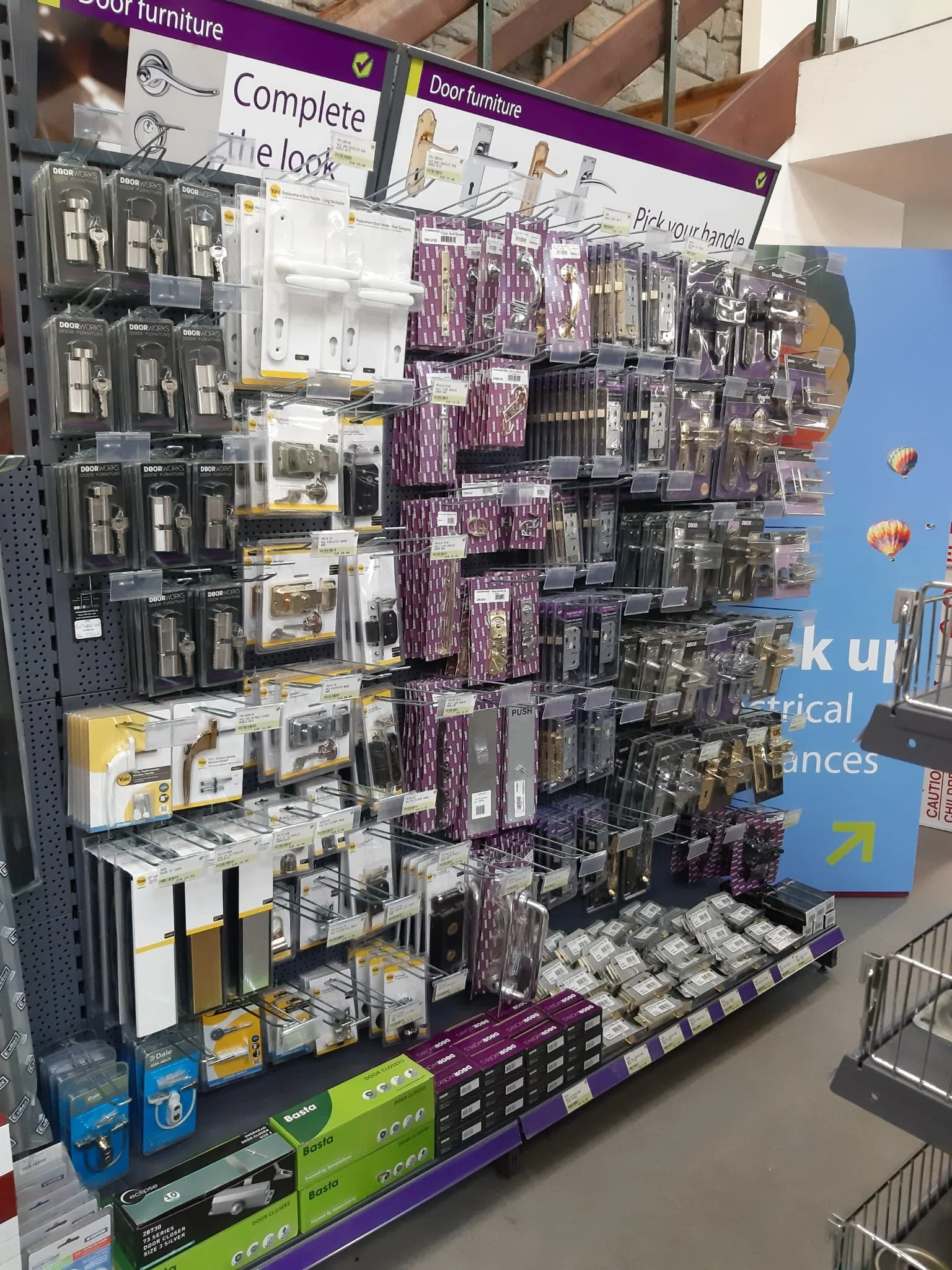 Store aisle with a variety of door hardware, including locks, handles, hinges, and door closers on display shelves. There are purple and white signs indicating the section for door furniture.