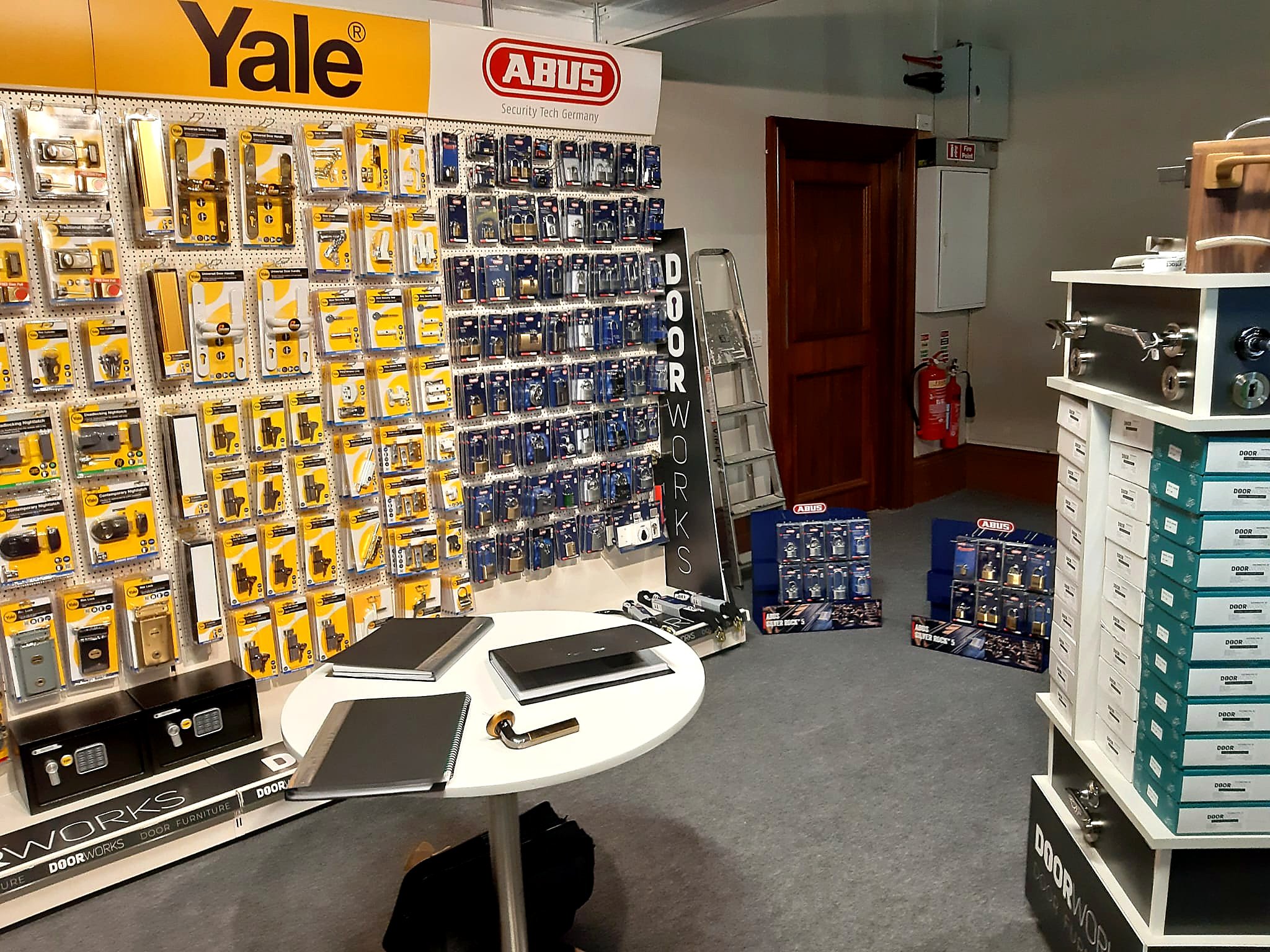 View inside a hardware store with shelves displaying locks, door handles, and security accessories. A round white table with notebooks, a lock, and electronic devices is also visible.