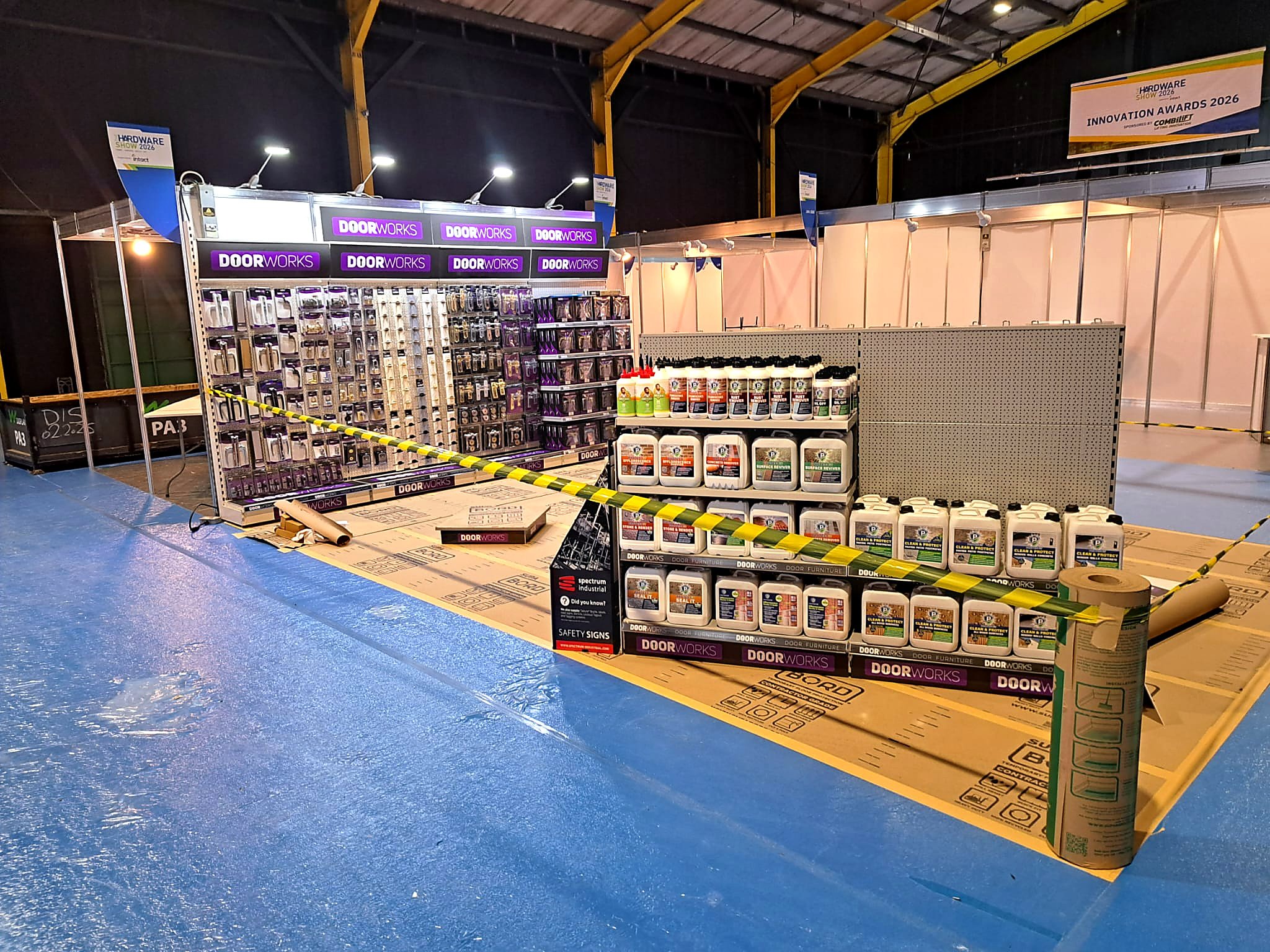 Trade show booth with shelves displaying door hardware and cleaning products, signs for 'DoorWorks' and 'Innovation Awards 2026,' safety caution tape on the floor and cardboard packaging, with a dark industrial ceiling overhead.