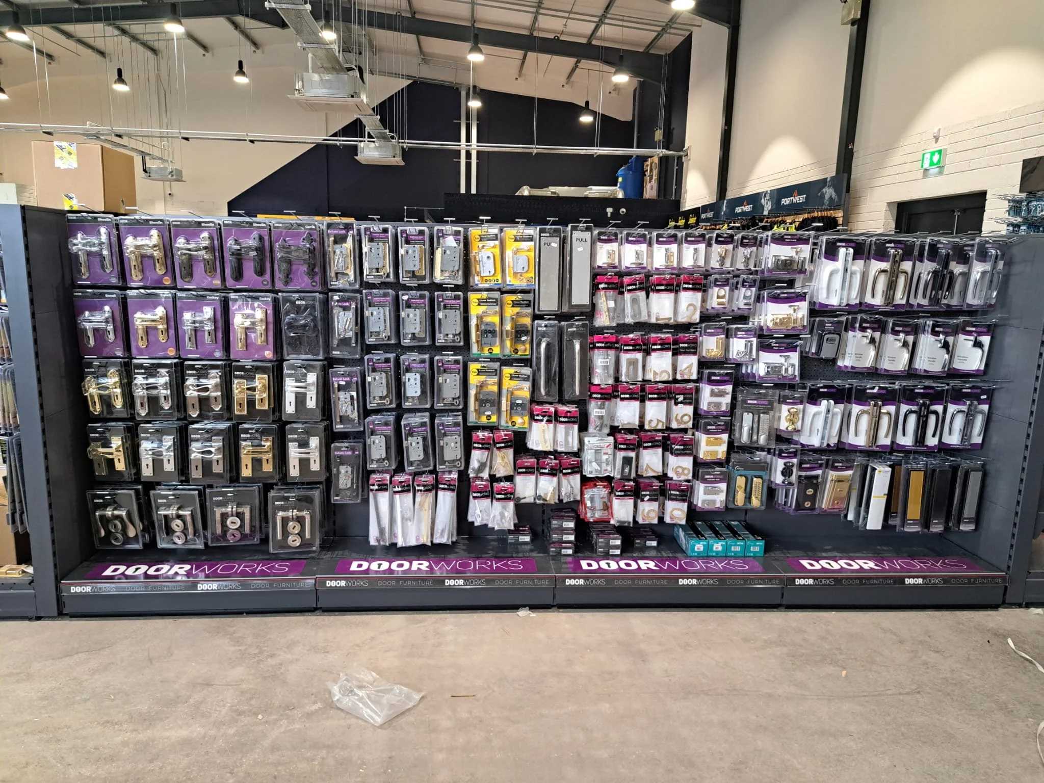 Display of door hardware such as locks and handles in a retail store aisle, with a black display rack and purple branding. The store has an industrial ceiling with overhead lighting.