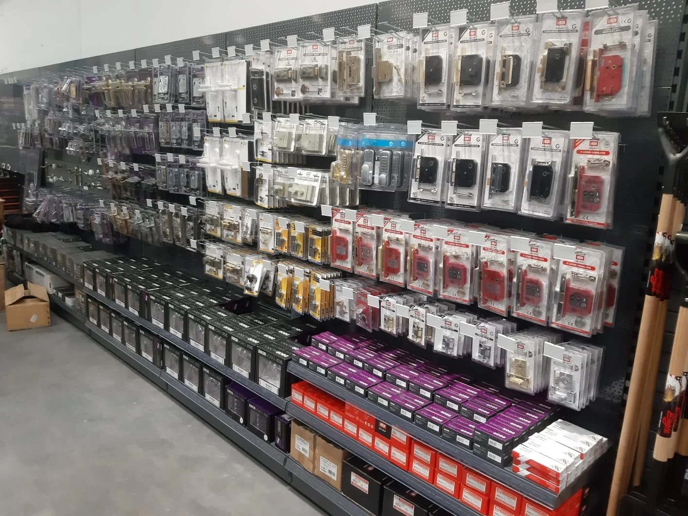 Retail store aisle with lock and latch hardware on display, including various types of locks, keypads, and related accessories arranged on a black pegboard wall and shelves.