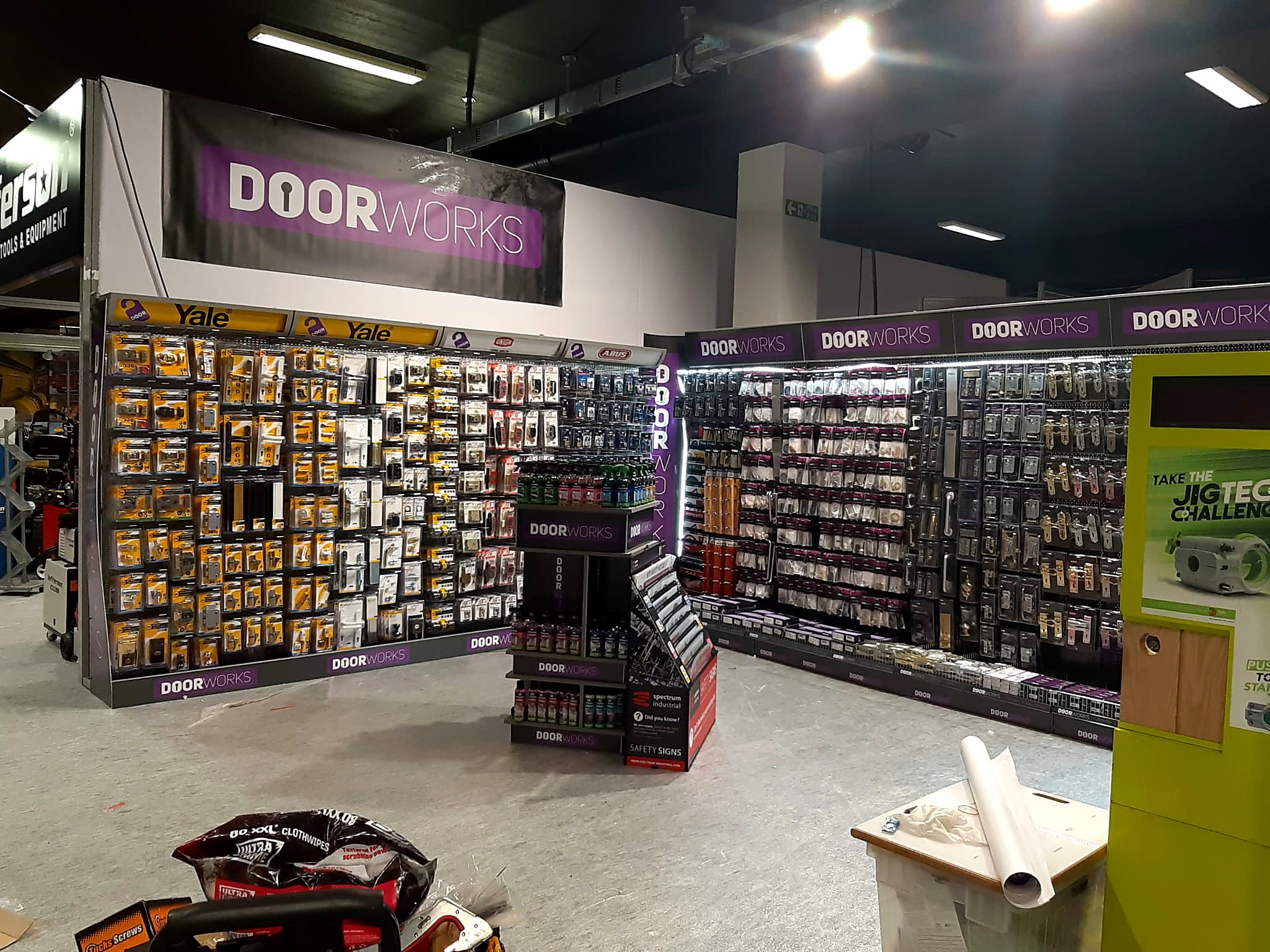 A retail store aisle displays door hardware including locks and handles on black pegboard panels with purple and white 'Door Works' branding. There is a yellow sign for Yale brand locks and various tools and safety signs on the floor.