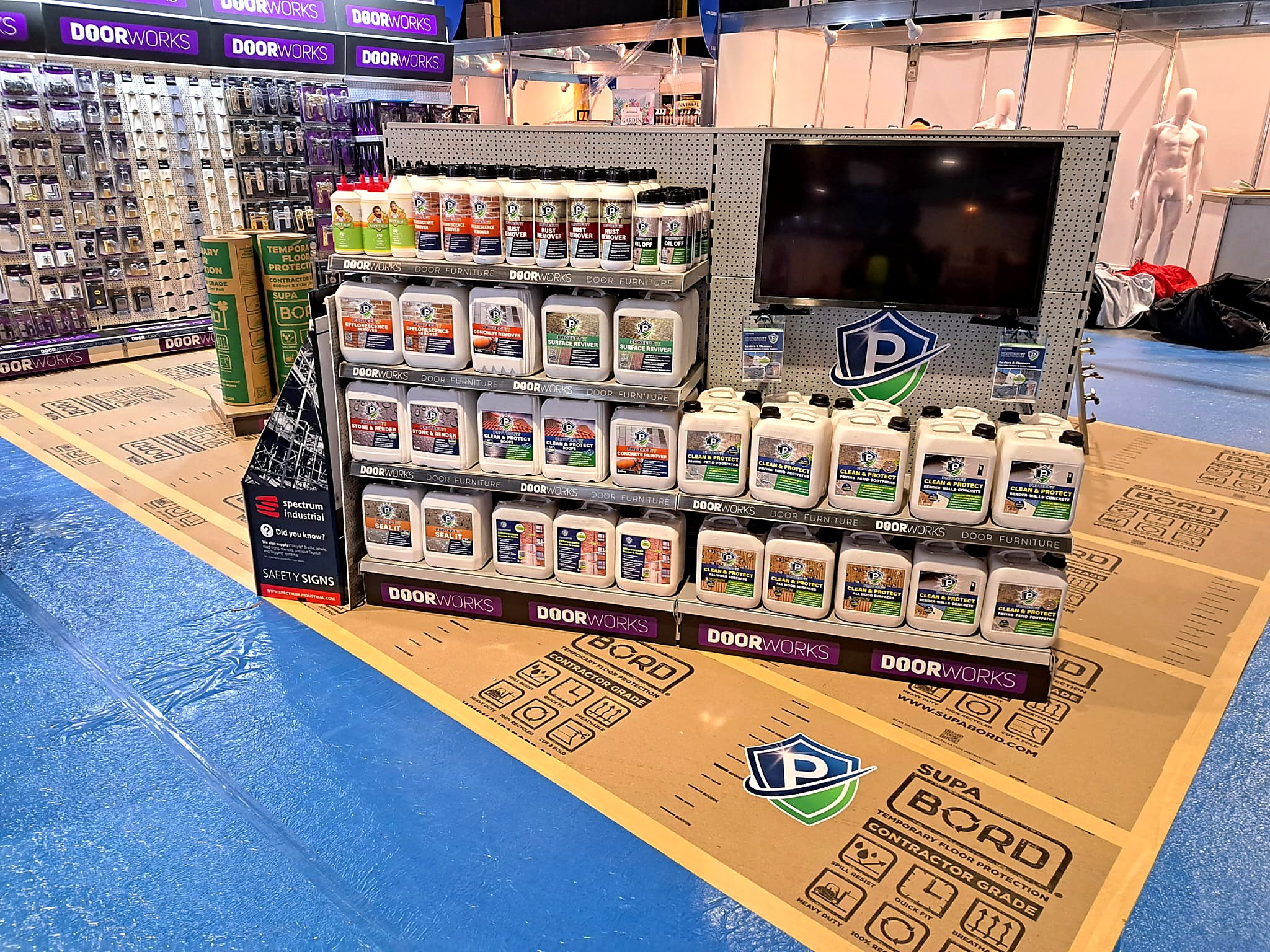 A display of cleaning products and supplies at a hardware store, including bottles and containers of surface remover, concrete remover, and other cleaning solutions, with a large TV screen and mannequins in the background.