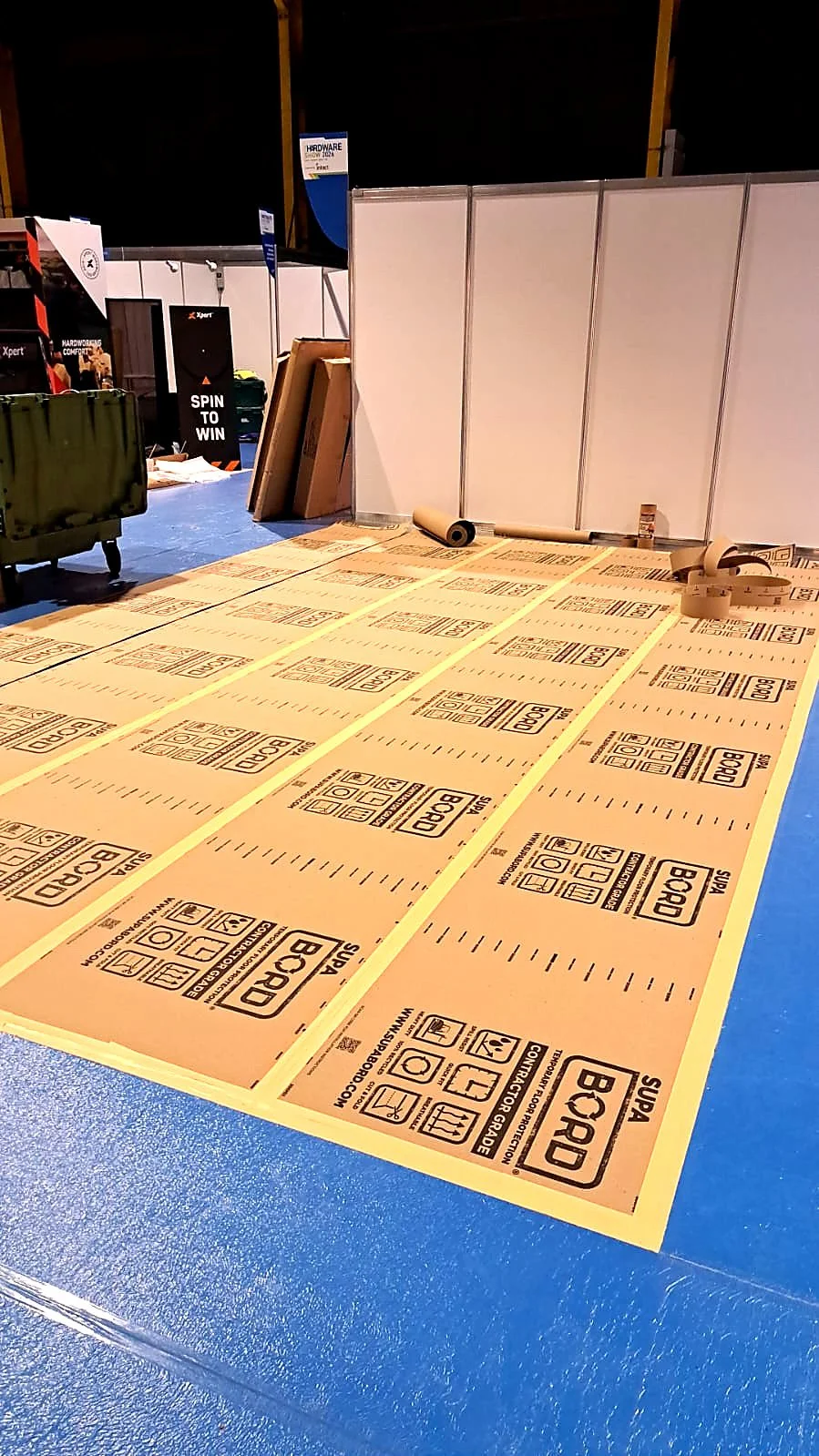 Construction floor with printed cardboard panels, yellow tape lines, in an exhibition or trade show booth area.