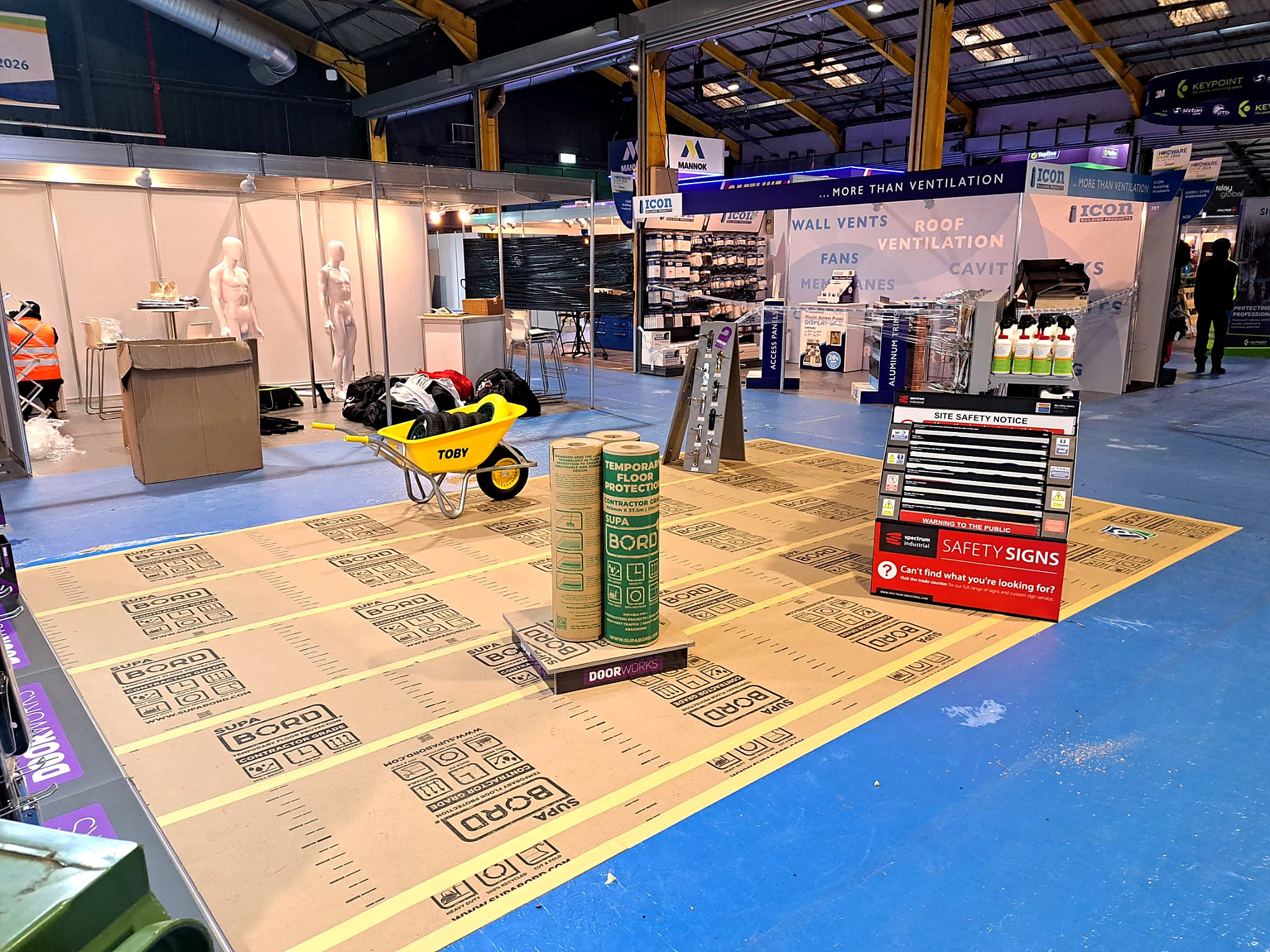 Construction zone with safety flooring, safety signs, and various building materials in an indoor setting, likely at a trade show or construction site exhibit.