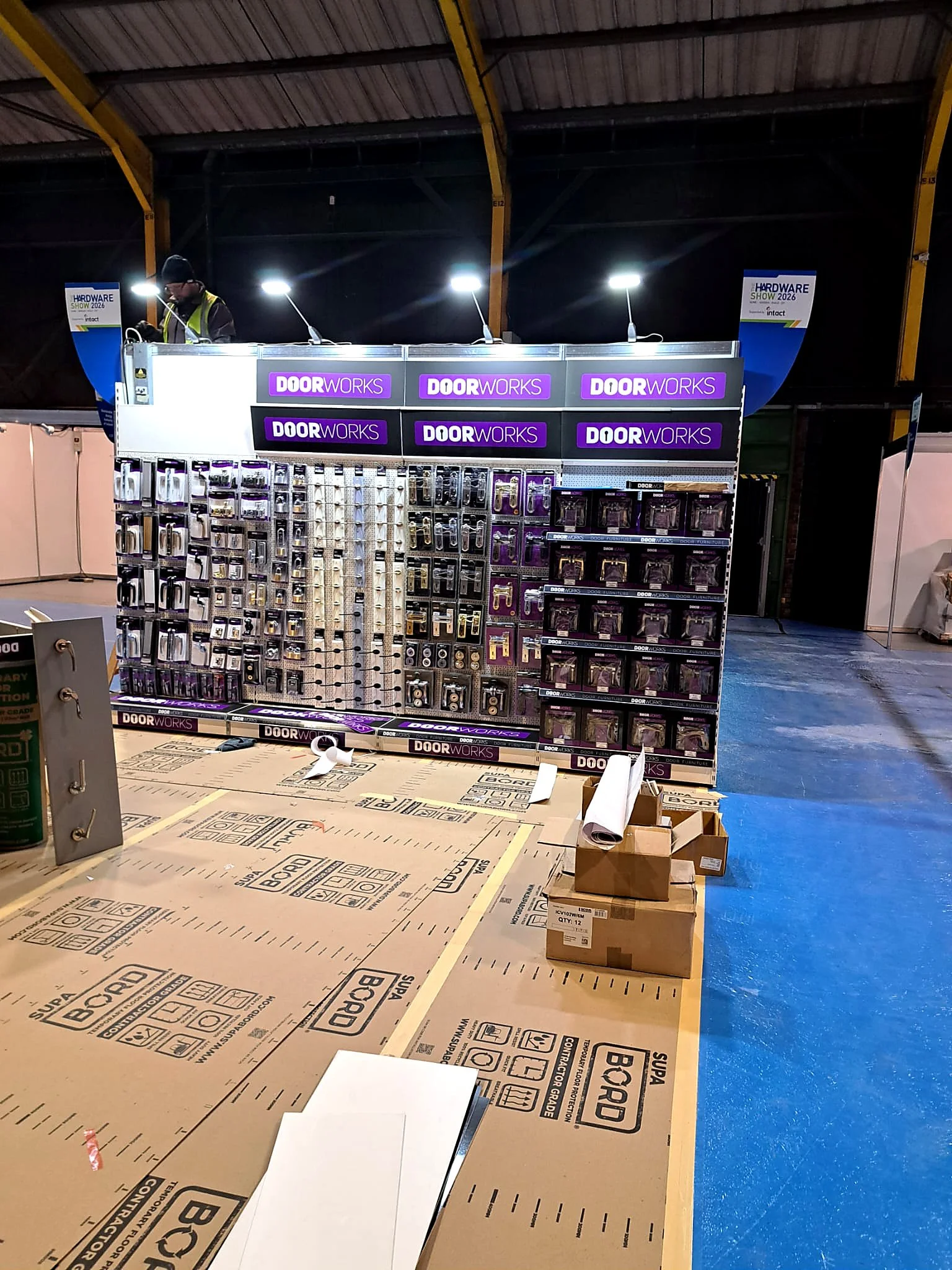 A person in a yellow safety vest and black hat working on a display of door hardware items at the Hardware Show 2023. The display features various door handles, locks, and accessories, with purple and black branding labeled 'DoorWorks'. The exhibitio