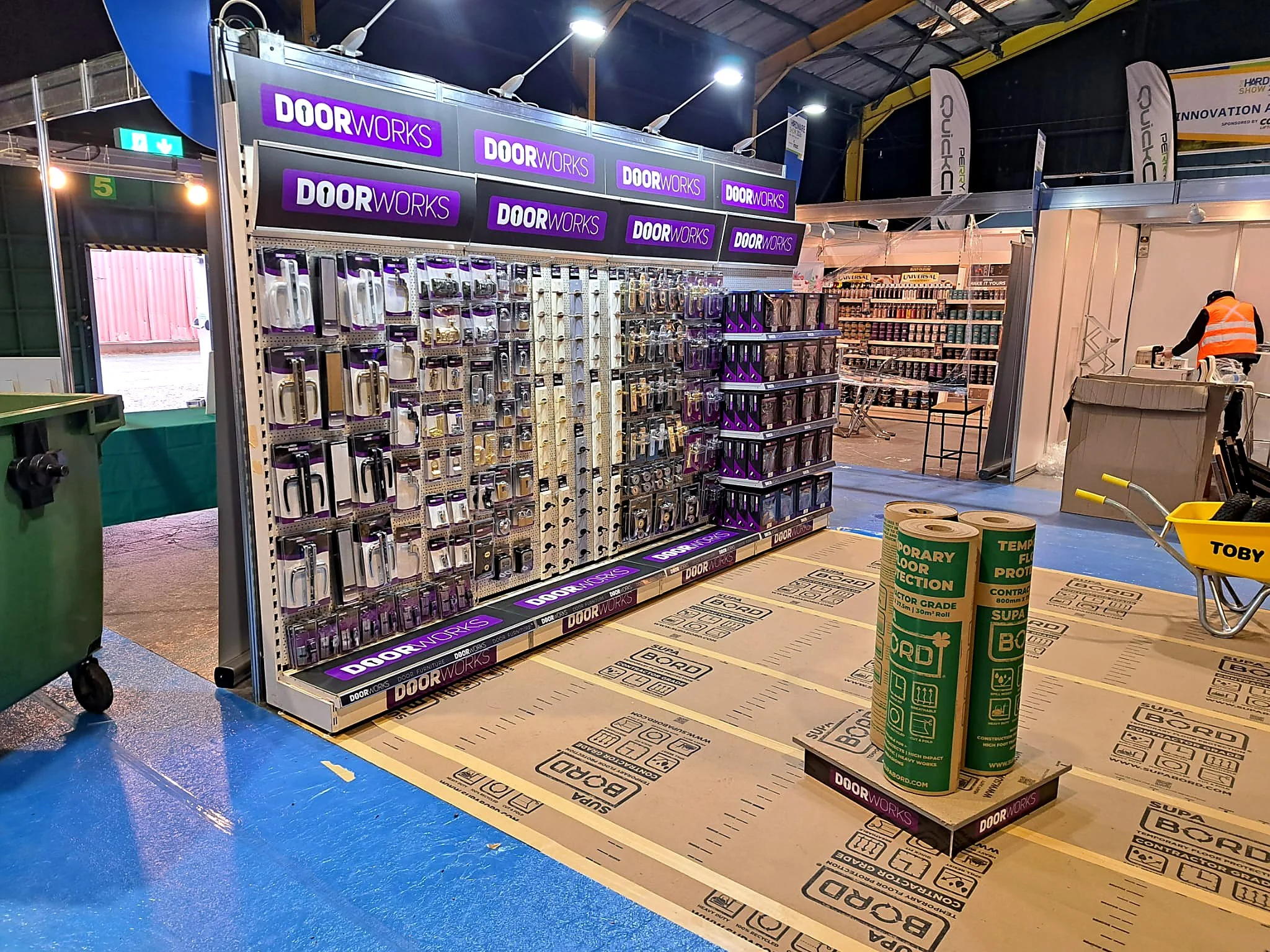 Store display with locks and hardware accessories branded 'DoorWorks,' bright overhead lighting, a person in a reflective vest, and various tools and packages on the shelves.