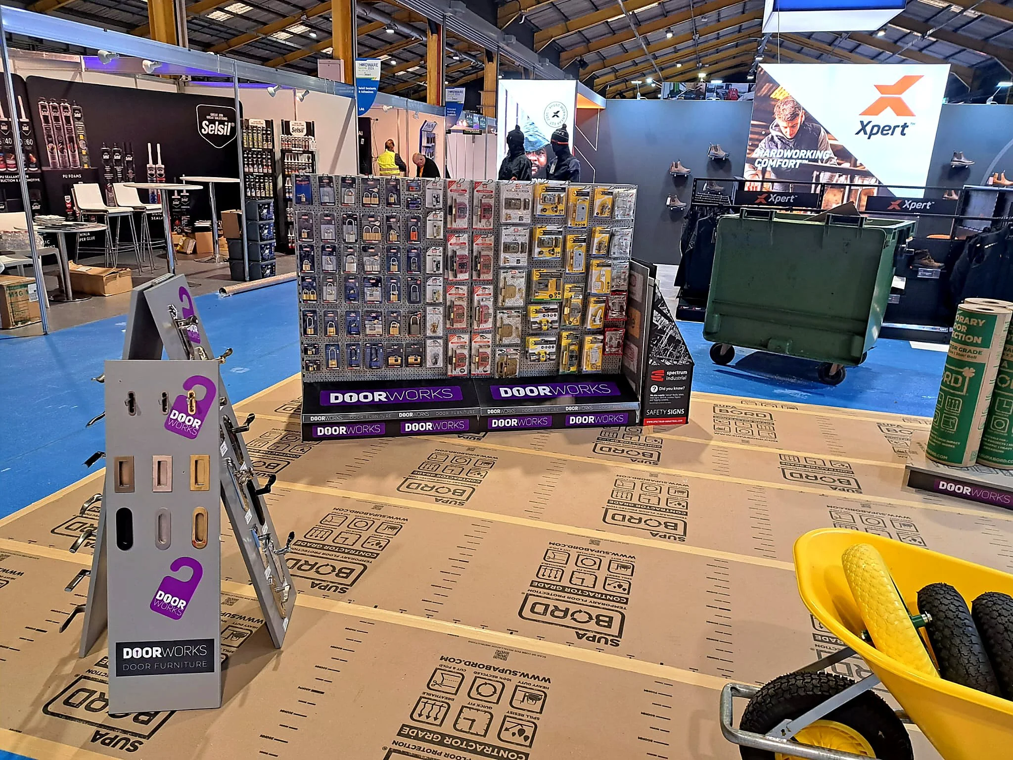 Trade show inside a large warehouse with booths displaying hardware supplies, a yellow wheelbarrow with black rocks and yellow gravel in the foreground, and various signs and promotional displays for door and hardware brands.
