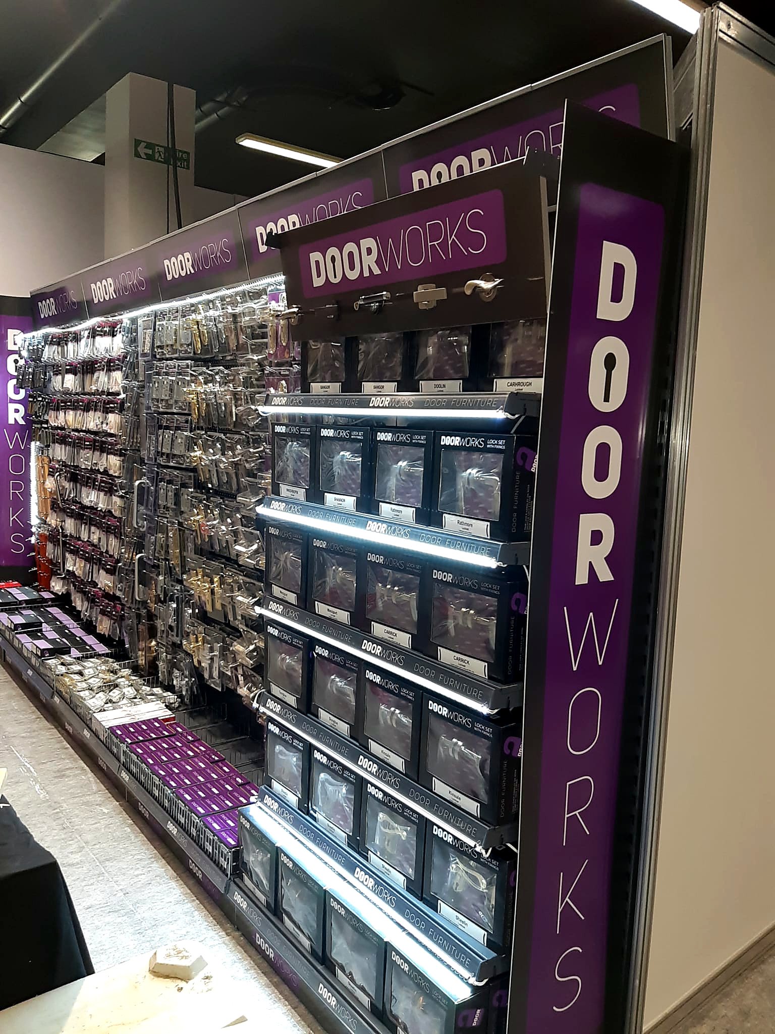 Display of door hardware and locks at a store, with various types of handles and lock sets arranged on shelves, and a purple and black sign reading 'DOORWORKS'.