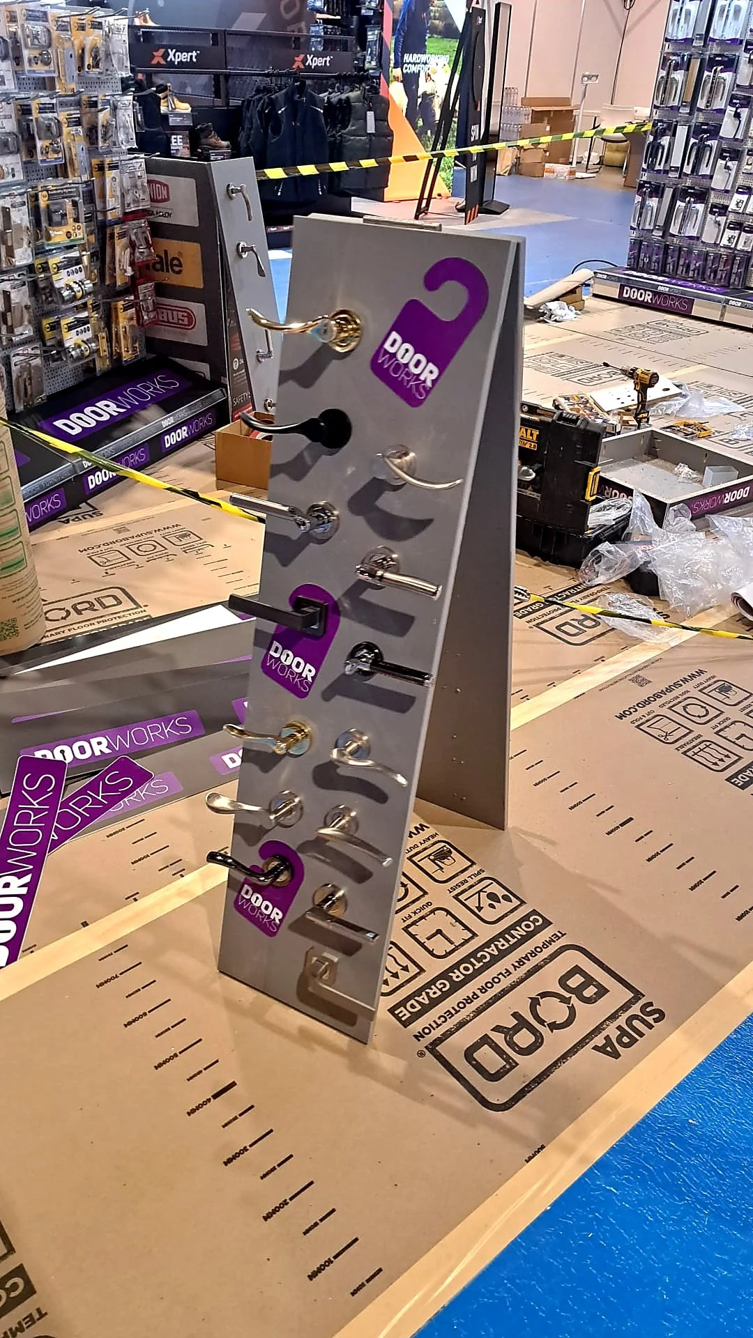 Display stand with various door hardware samples at a hardware store, with the store's signage and shelves in the background.