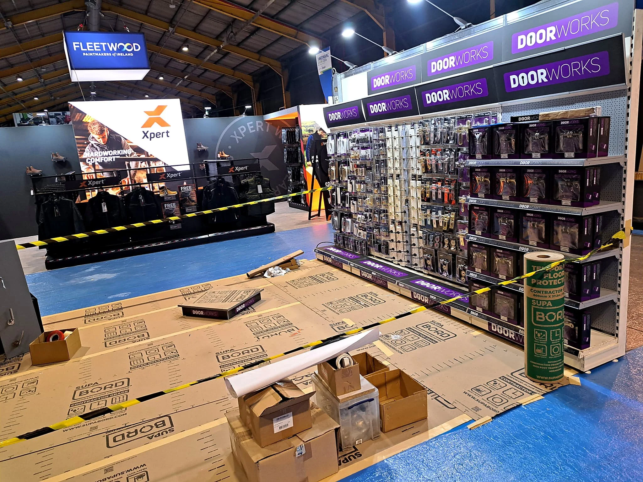 Inside a store or warehouse, with shelves stocked with door handles and hardware, packaging materials, and large boxes on the floor. Multiple signs and posters are visible, including a large digital screen displaying "FLEETWOOD PAINTMAKERS OF IRELAND