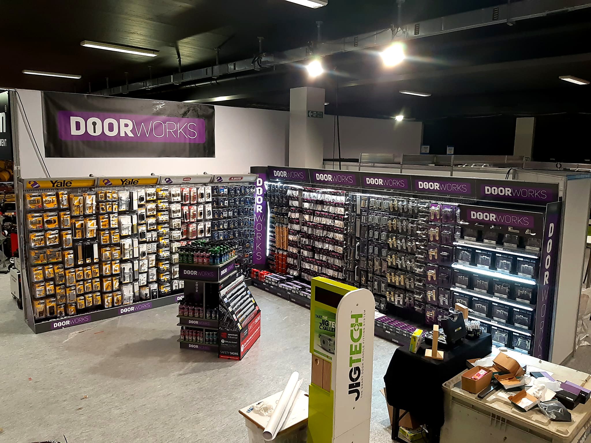 Inside a hardware store with shelves stocked with door hardware and accessories. There are multiple purple and white signs reading 'DOOR WORKS.' Various products are displayed on the shelves, and the floor appears to be concrete. The store has a dark