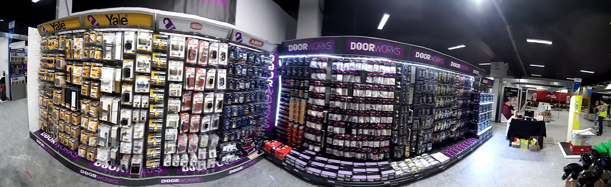 A store aisle displaying door hardware accessories and supplies on shelves, with a worker in the background wearing a yellow safety vest.