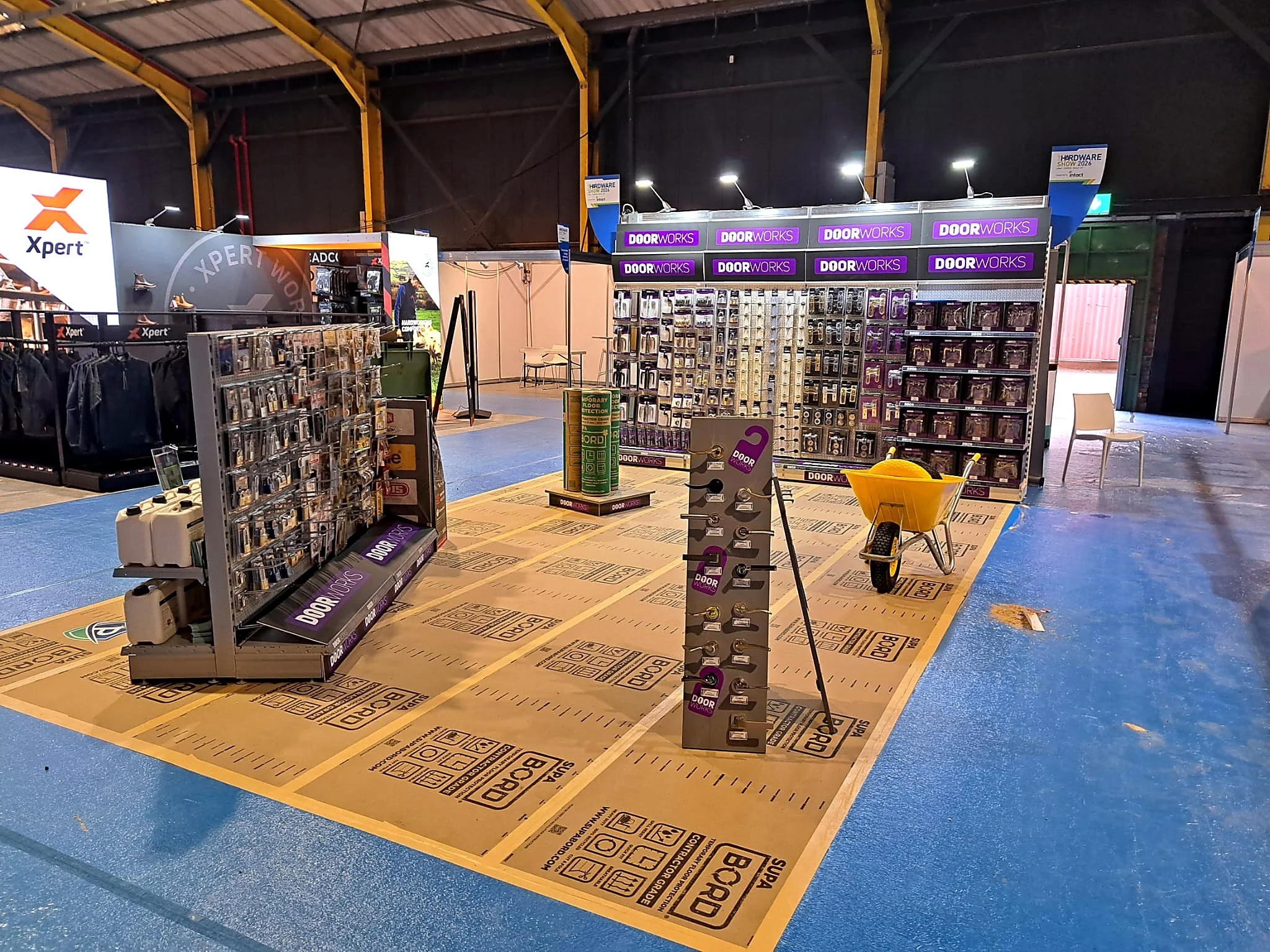 A hardware store display area with various lock and door handle samples, a large Xpert sign, purple DoorWorks signage, a yellow shopping cart, and a section for subwoofer installation. The floor shows Paper and BOPD markings.