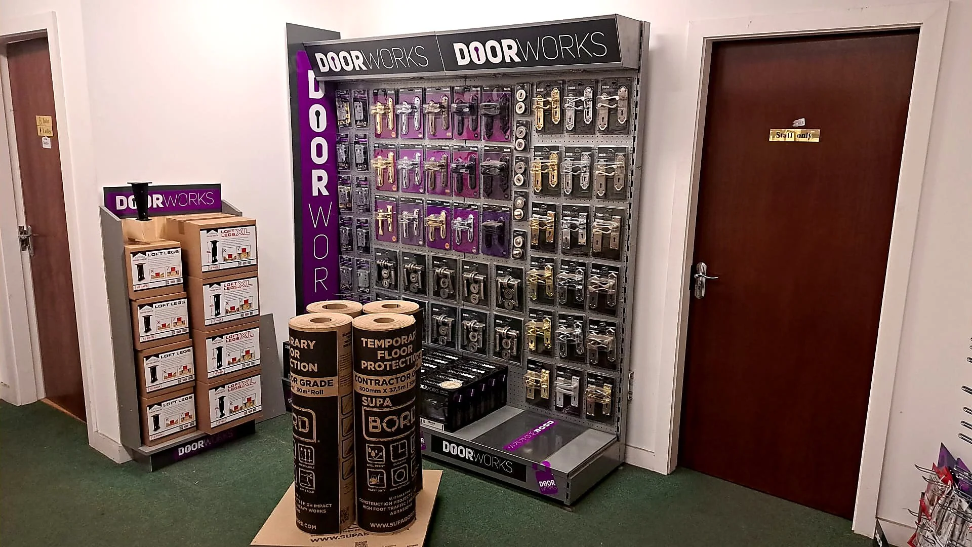 Display of various door handles and locks at a hardware store, with packaging and labels, alongside rolls of floor protection material.
