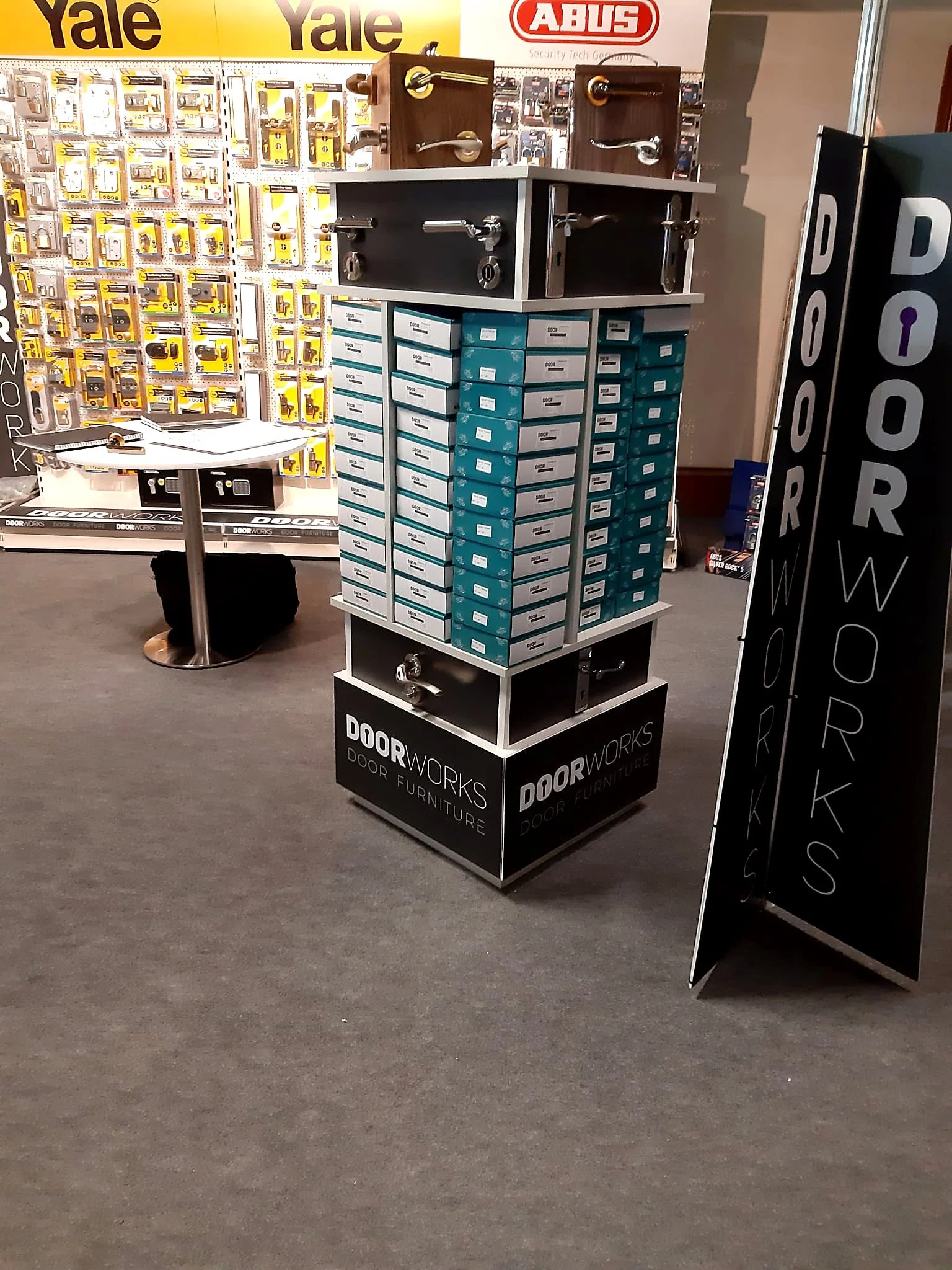 Display booth for door hardware and furniture at a trade show with locks, handles, and boxes of door hinges, with signage for Yale and ABUS brands.