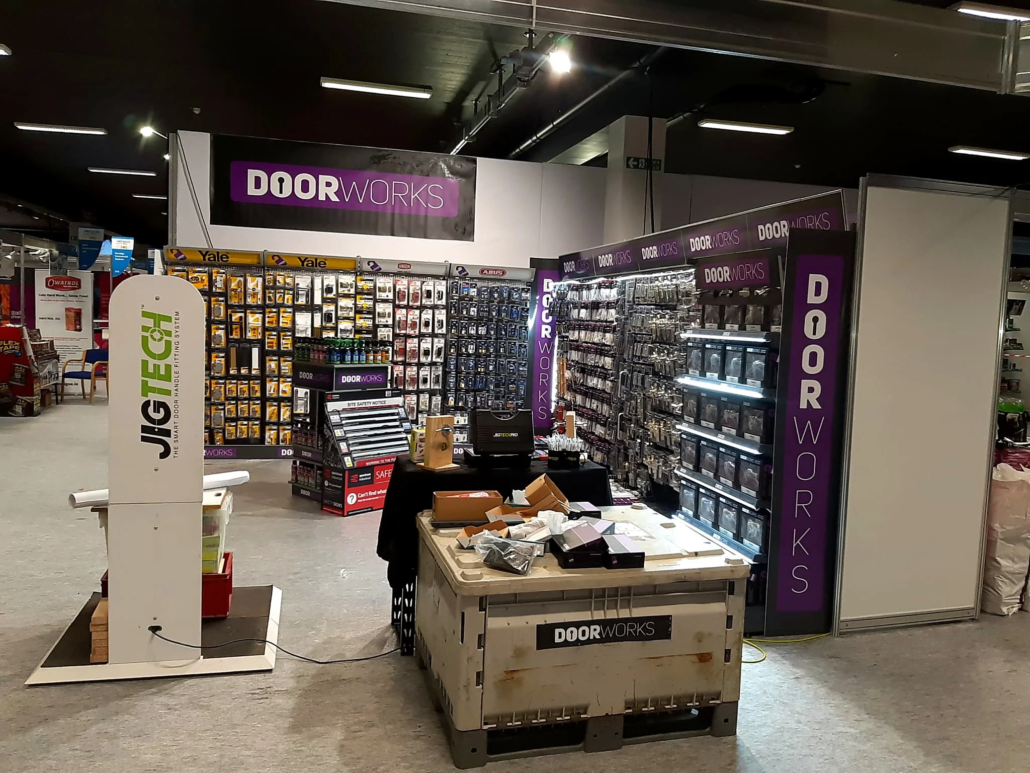 Display of door hardware and accessories in a store, with a large purple and black sign reading 'DOOR WORKS' in the background, and various tools and supplies arranged on shelves and tables.