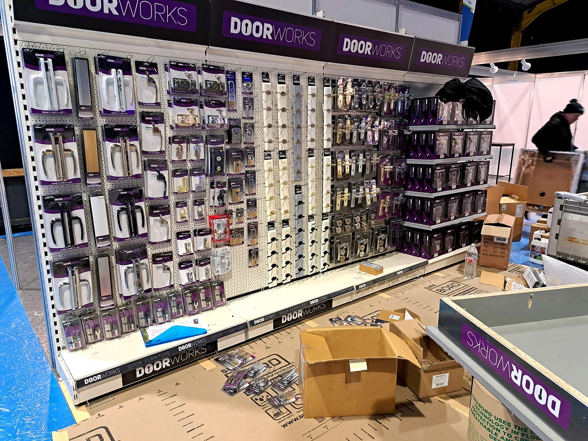 A retail display of door hardware and accessories arranged on pegboard and shelves, including various door handles, locks, and hardware with purple and black branding for DOORWORKS.