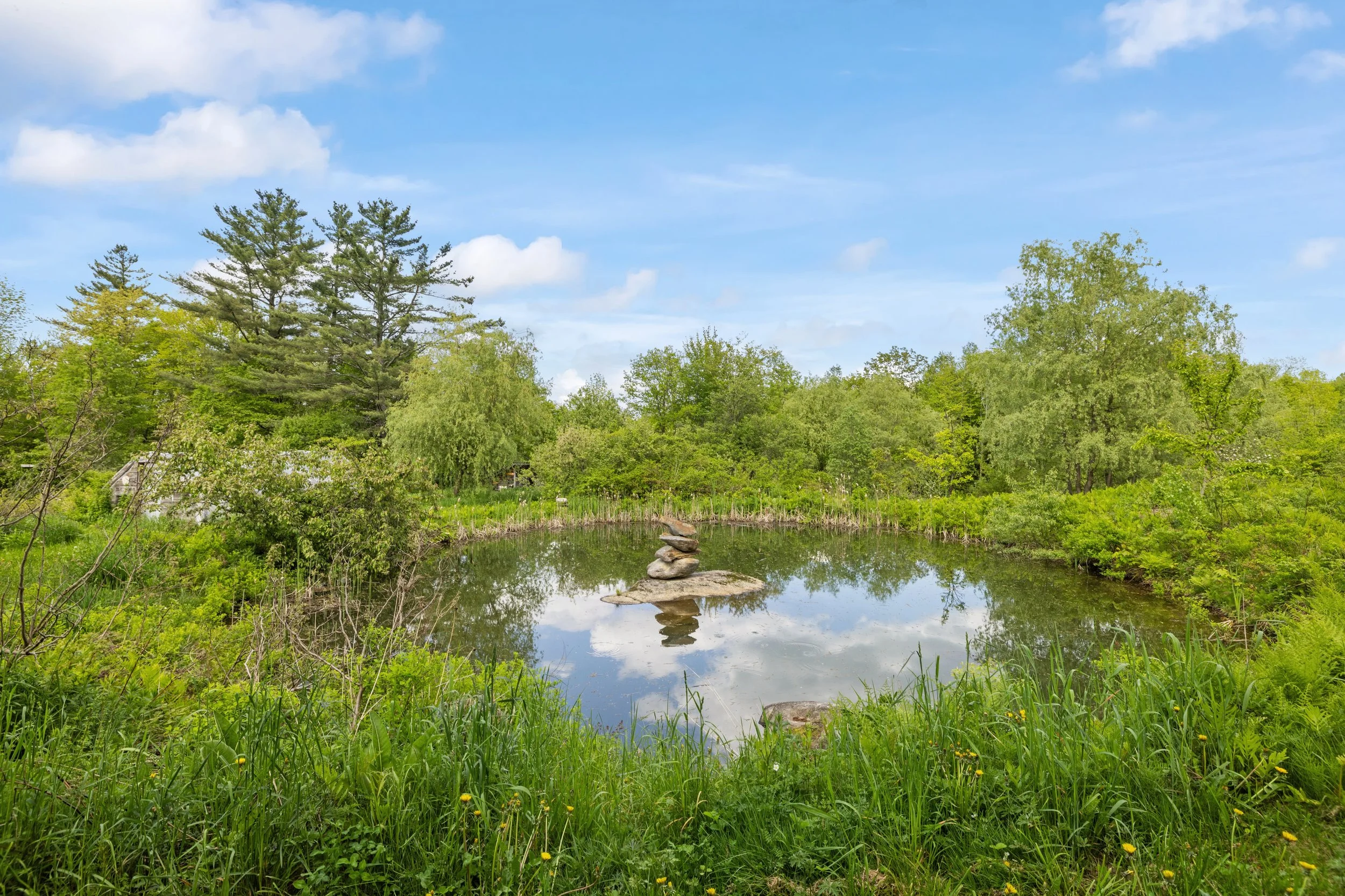 A small pond surrounded by lush green trees with a stack of rocks on a flat stone in the center of the pond, and a blue sky with a few clouds reflected in the water.