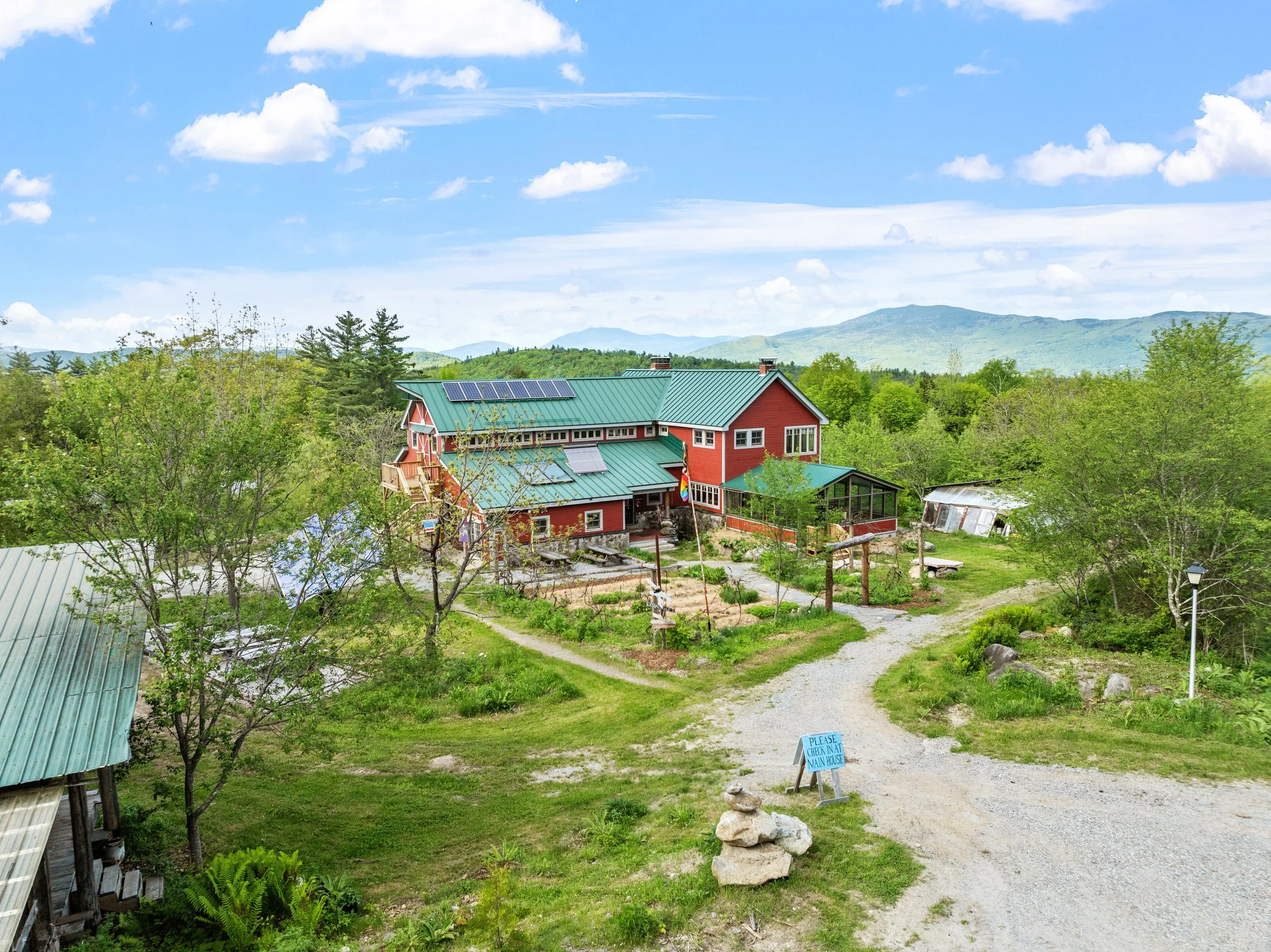A red house with a green metal roof and solar panels, surrounded by lush green trees, with a gravel pathway leading up to it and mountains in the distance under a partly cloudy sky.