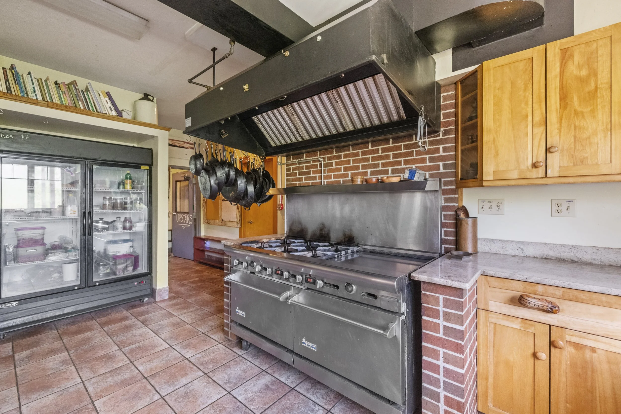 Commercial kitchen with a large stove, hanging pots, wooden cabinets, and glass door refrigerator.