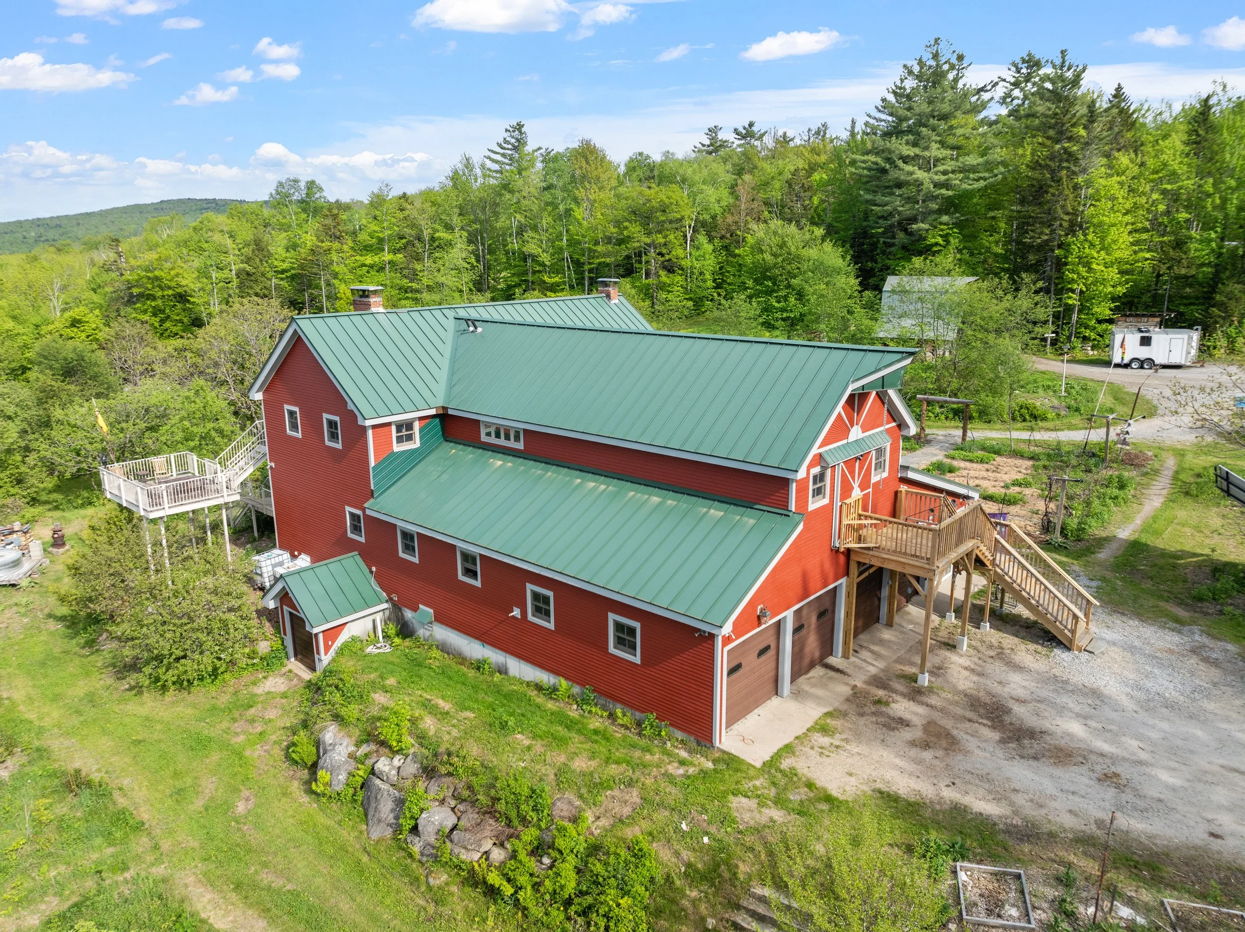 Aerial view of a large red barn with a green metal roof, surrounded by green trees and grass, with a gravel driveway and a trailer in the background.