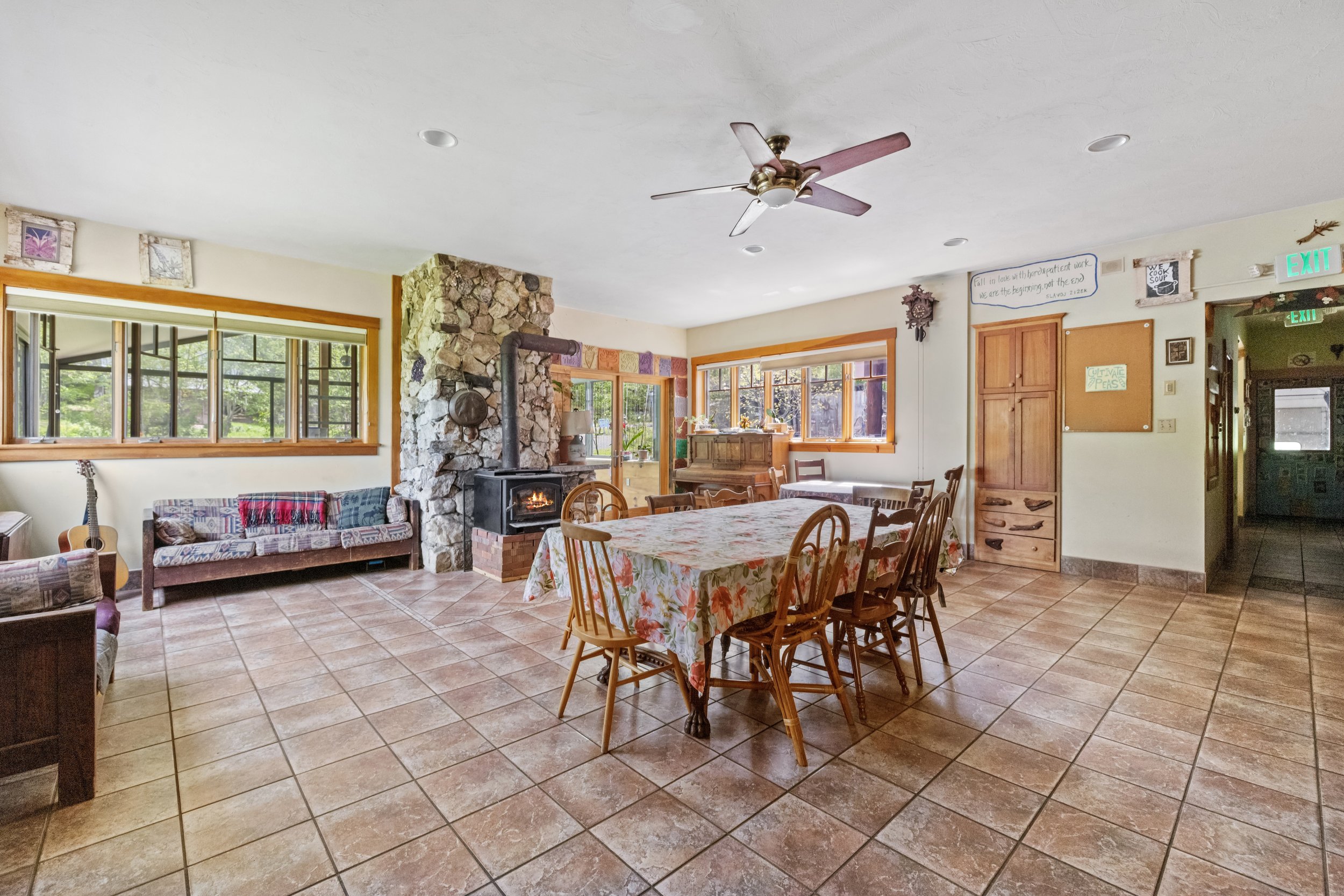 A spacious dining area with a large rectangular table covered with a floral tablecloth, surrounded by wooden chairs, and a fireplace with a stone chimney on the back wall. The room features large windows, tiled flooring, and a ceiling fan, with additional furniture and decorations, including a couch, a guitar, and a wooden cabinet.