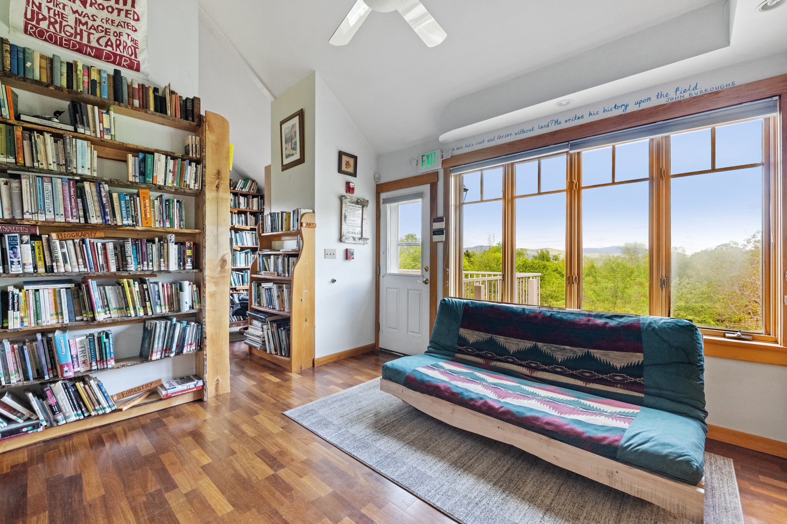 Cozy room with wooden flooring, large windows overlooking green trees, a wooden bookshelf filled with books, a cozy futon with a colorful cover, and wall decorations including a white sign, framed pictures, and a quote above the window.