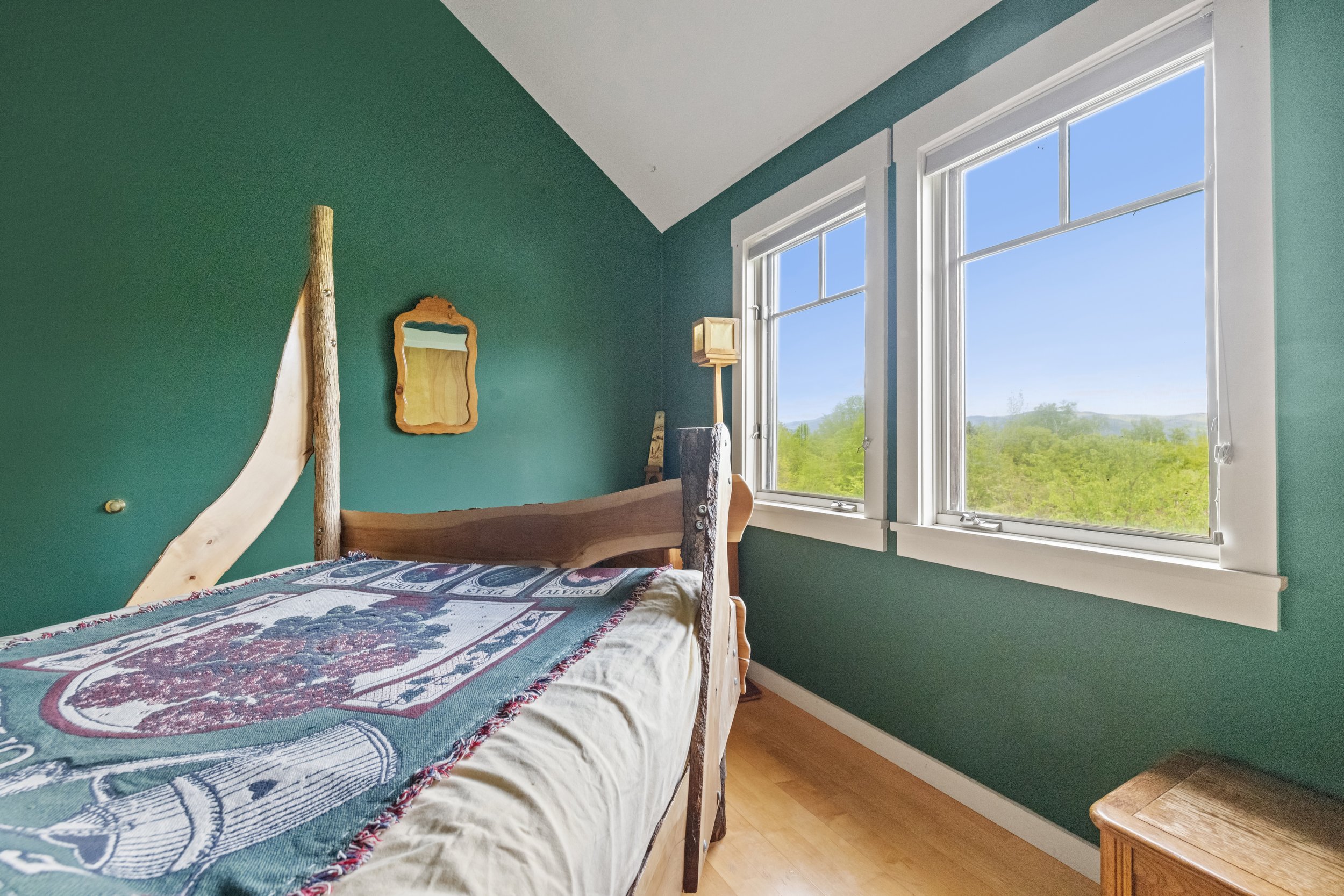 A bedroom with green walls, a wooden bed with a colorful blanket, two windows showing a green landscape outside, and a small wooden nightstand.