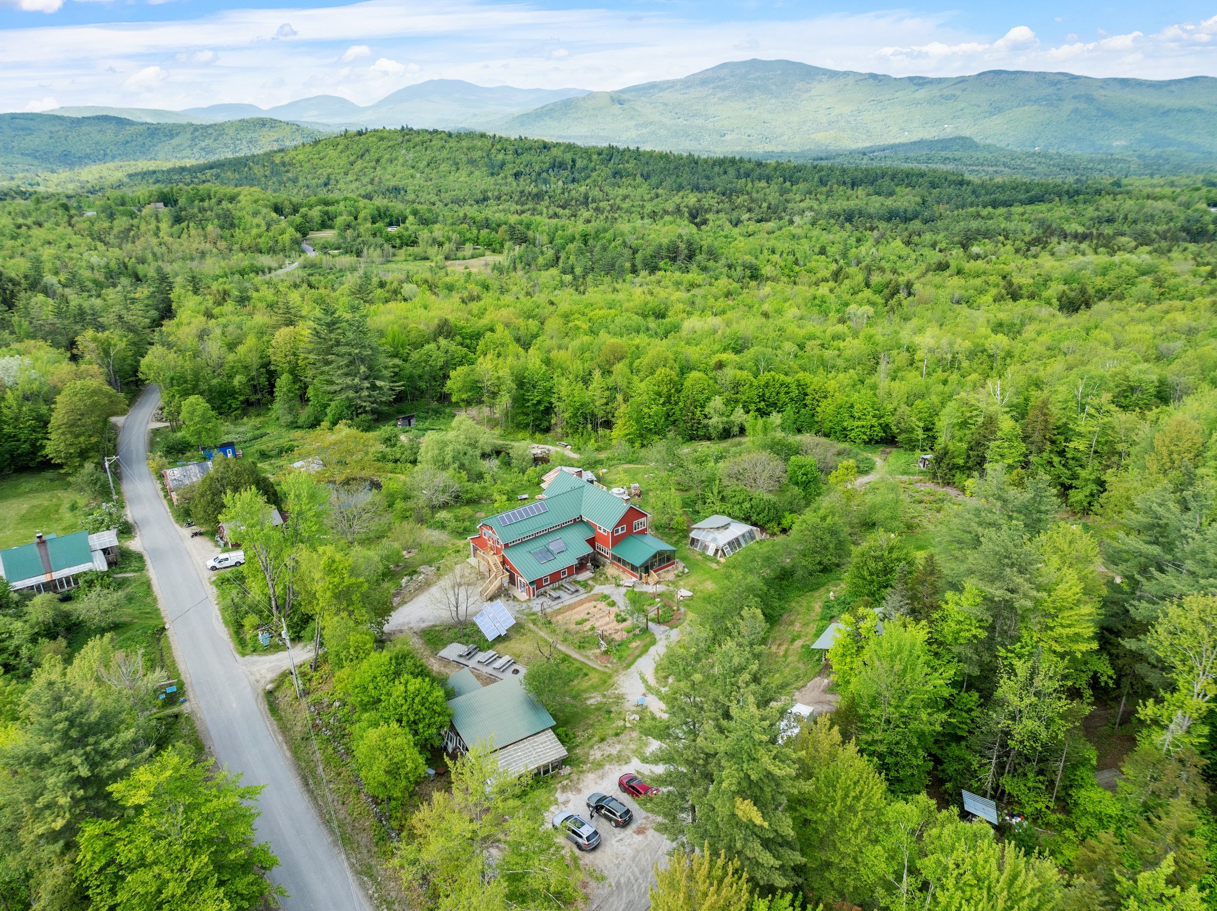 Aerial view of a rural landscape with a large red house with green roofing, surrounded by green trees, smaller houses, a road, cars, and distant rolling hills.