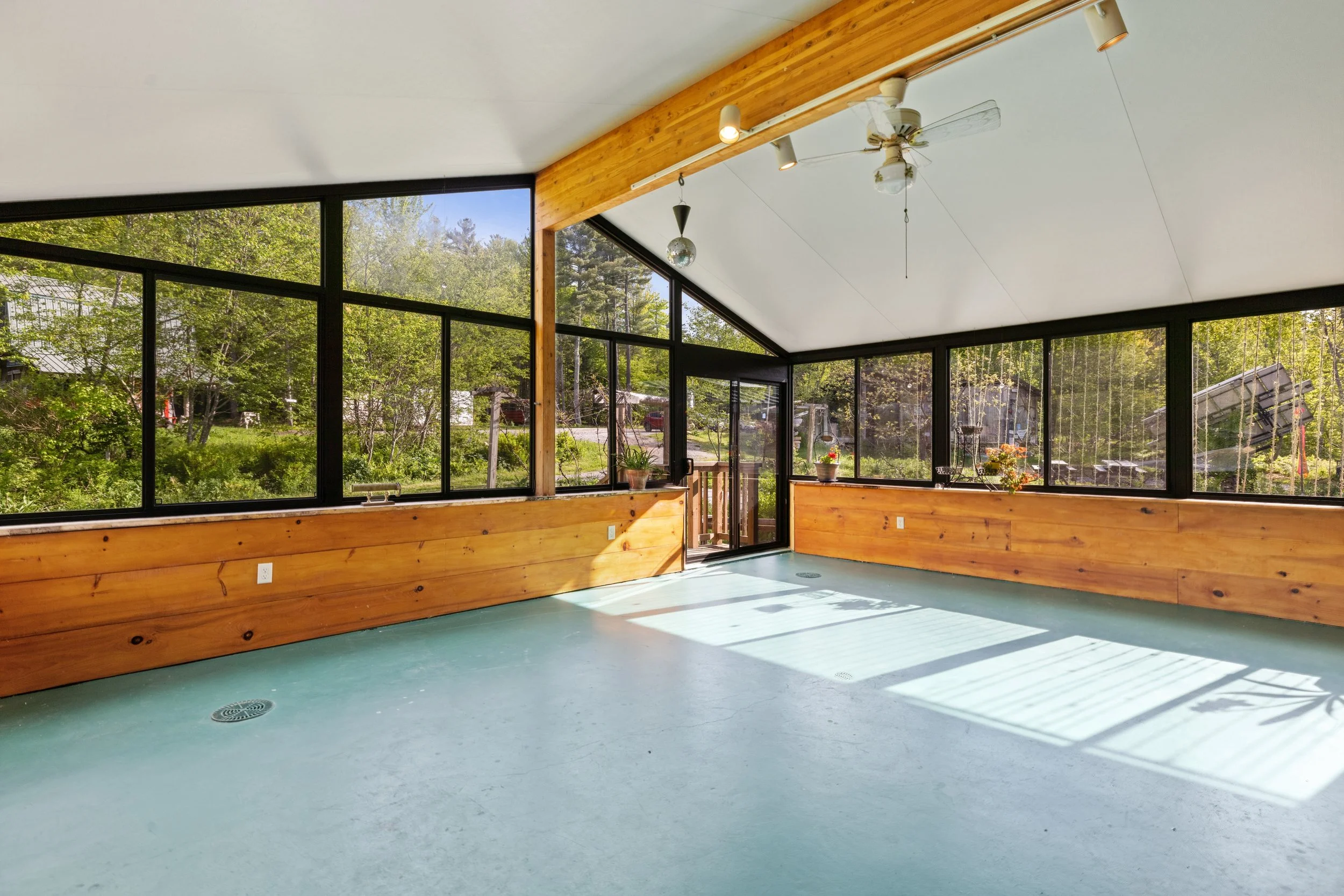Sunlit screened porch with large black-framed windows, wooden wall paneling, and a ceiling fan