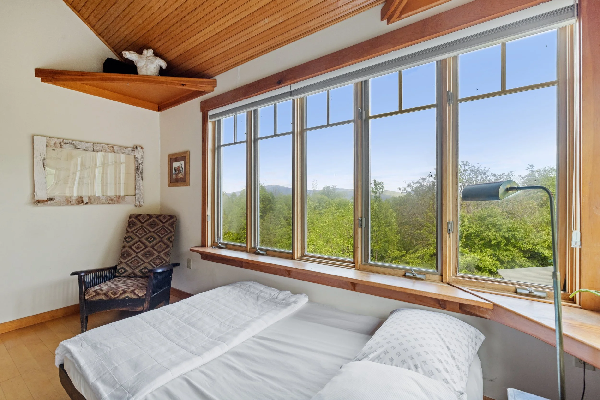 A bedroom with a large window showing trees outside, a bed with white bedding, a patterned armchair, a small wall mirror, framed pictures on the wall, and a green floor lamp.